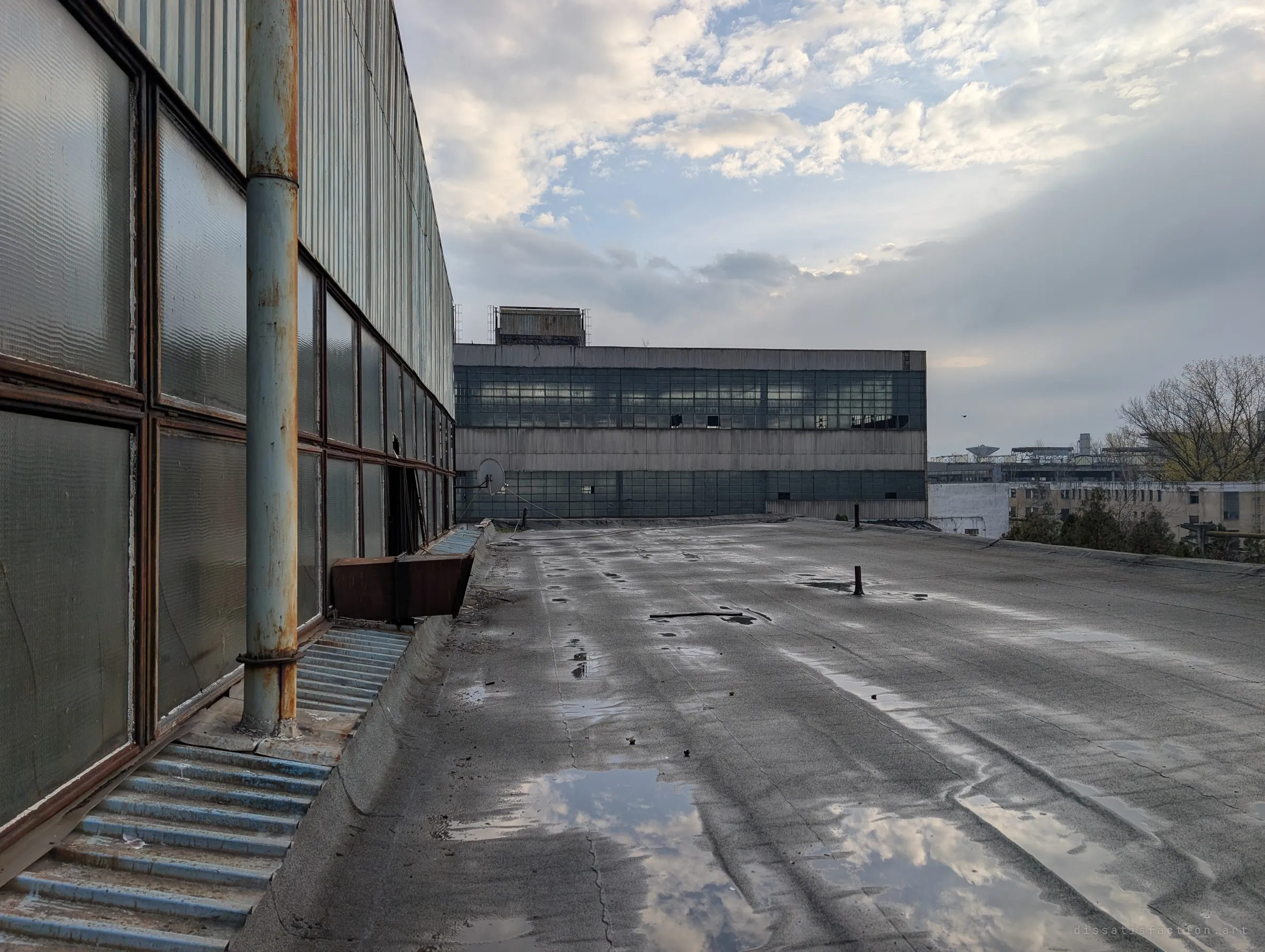 A flat puddled roof runs alongside a wall of ribbed glass panels and a rusting drainpipe toward a long glazed factory hall. Clouds reflect in the standing water.