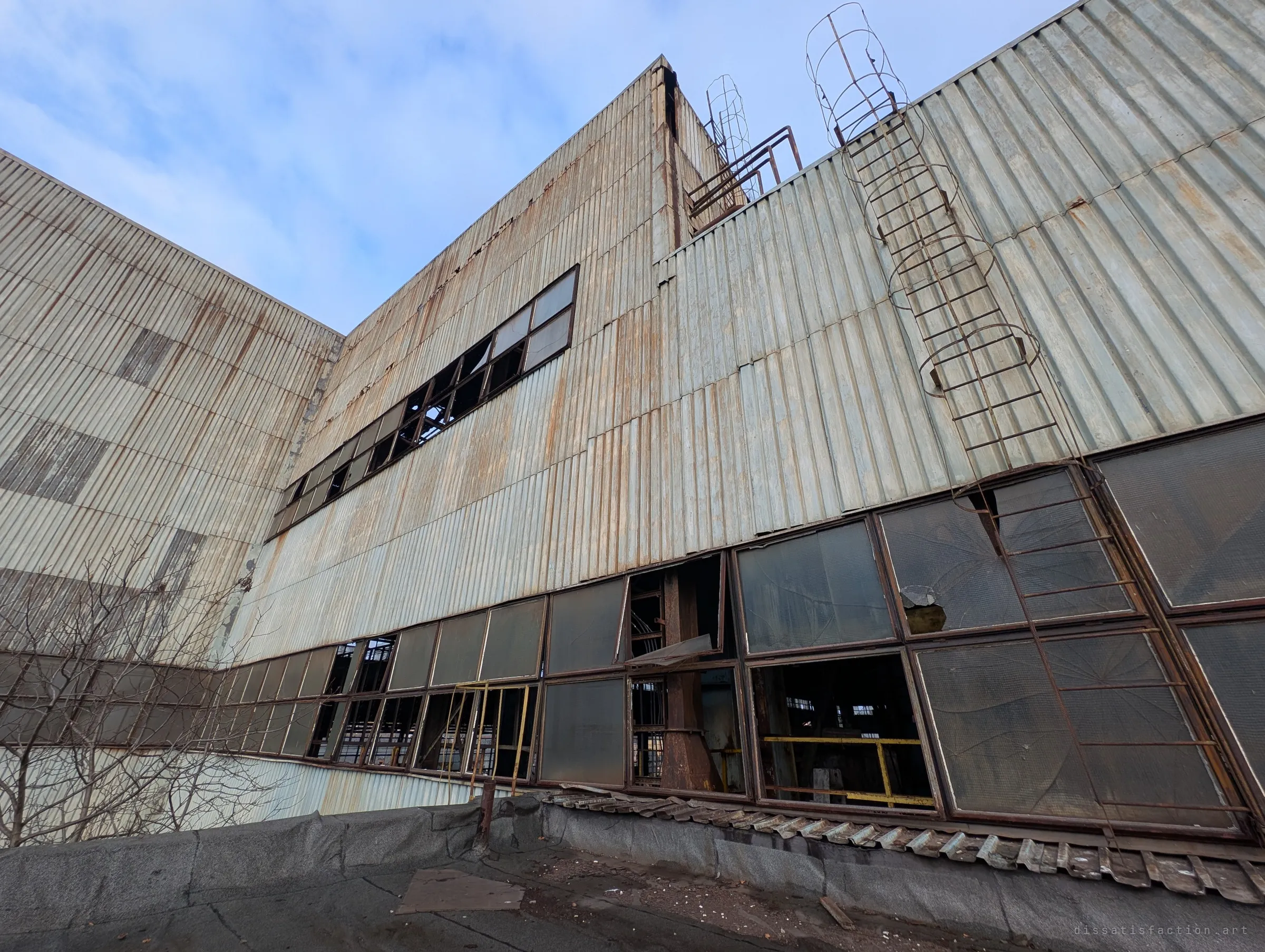 A low wide-angle view up a rust-streaked corrugated facade with a caged ladder and rows of broken windows. The scale of the hall towers over the roof it rises from.