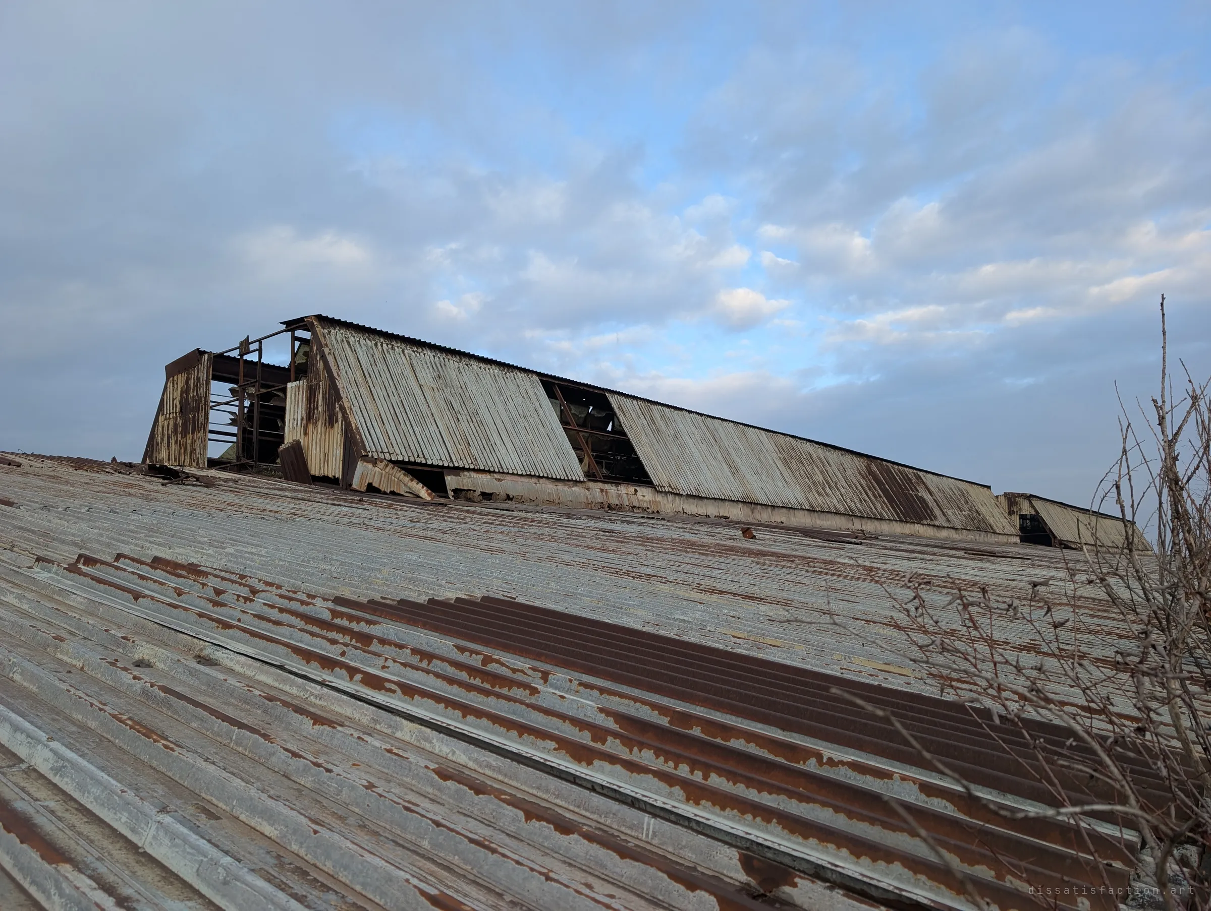 A slanted rusted corrugated roof leads up to a collapsing rooftop lantern with missing panels, bare branches creeping in from the right. The decay is visible in every sheet of metal.