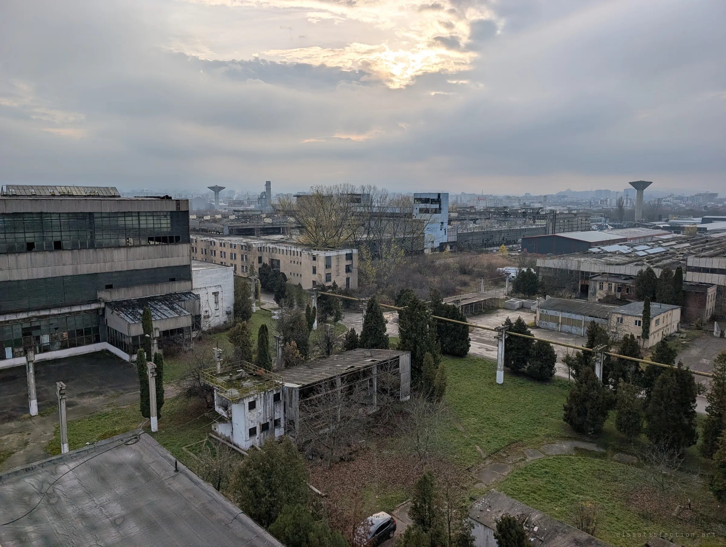 From a high rooftop, the abandoned works spreads out below: derelict halls, an overgrown courtyard crossed by pipe bridges, and cypress trees under a heavy sky. It shows how vast the site is.
