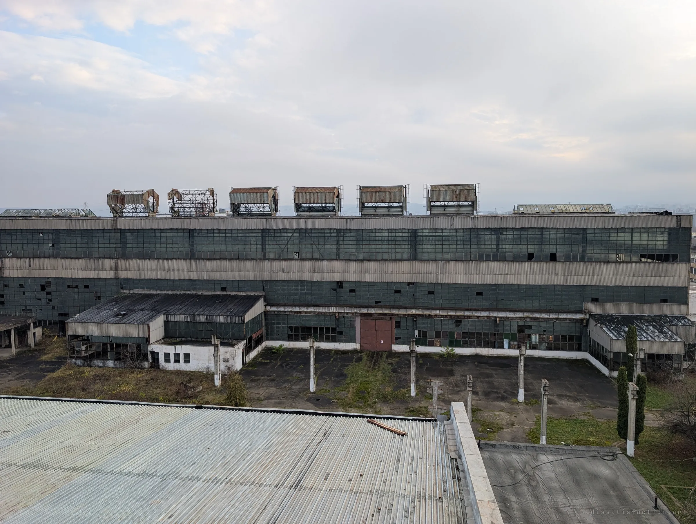 A huge glazed production hall stretches across the frame, its roof crowned by a row of rusting ventilator housings above an empty, weed-cracked yard. Most of its window panes are broken or missing.