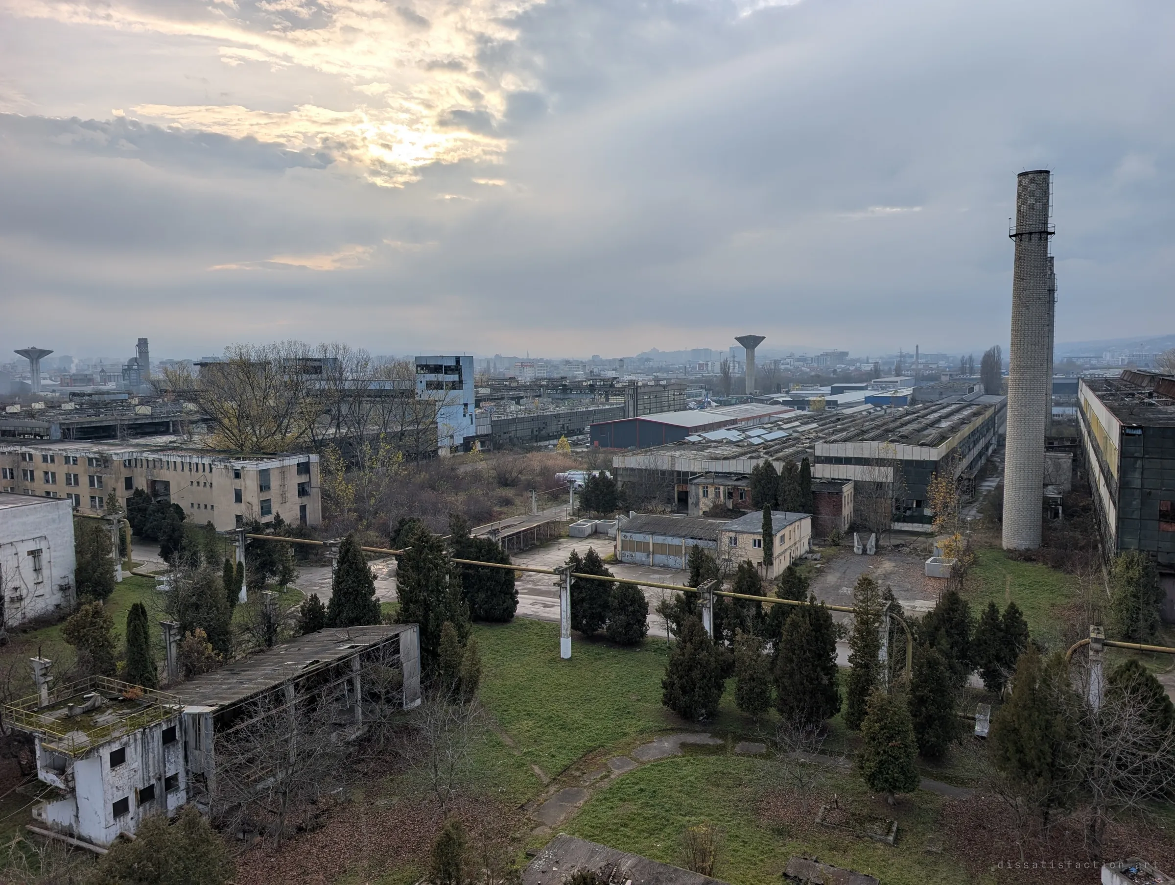 A high view over the overgrown grounds of the works, with a tall brick chimney on the right and rows of derelict halls fading into haze. Low sun breaks through the clouds.