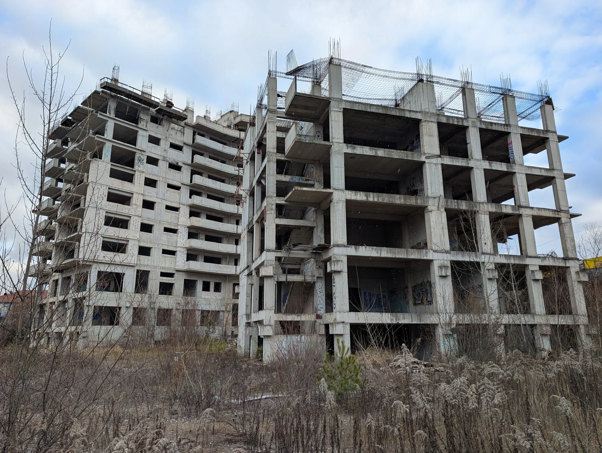 Two abandoned concrete tower skeletons rise above frosted weeds, rebar still sprouting from their unfinished roofs. The establishing view of the site.