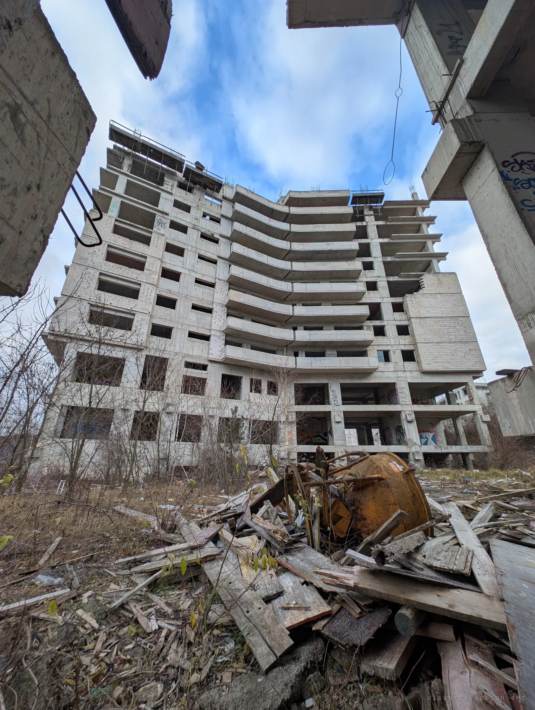 An ultrawide look up at the tallest block and its curved tiers of empty balconies, framed by concrete columns over a pile of broken timber and a rusted cement mixer.