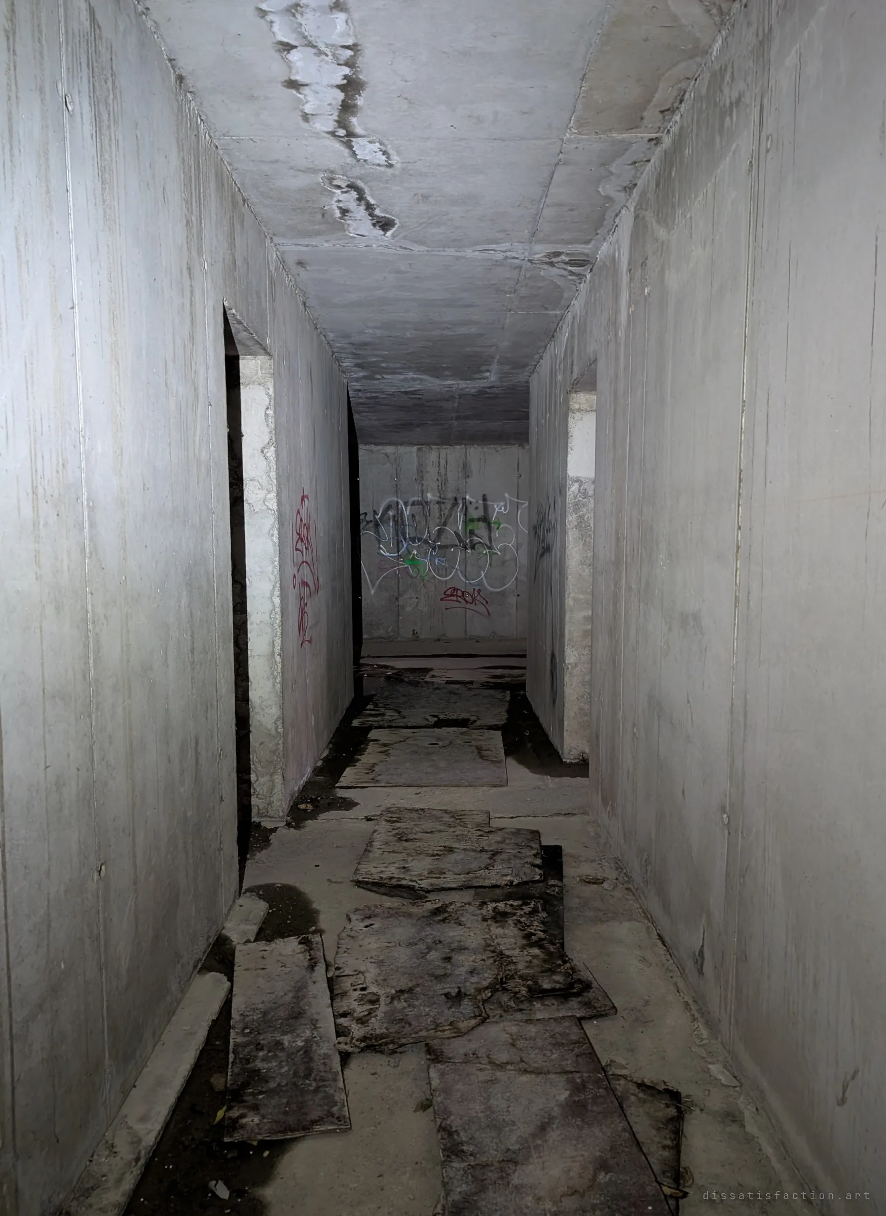 A dark bare-concrete corridor with loose boards laid over the wet floor, leading to a tagged wall at the far end.