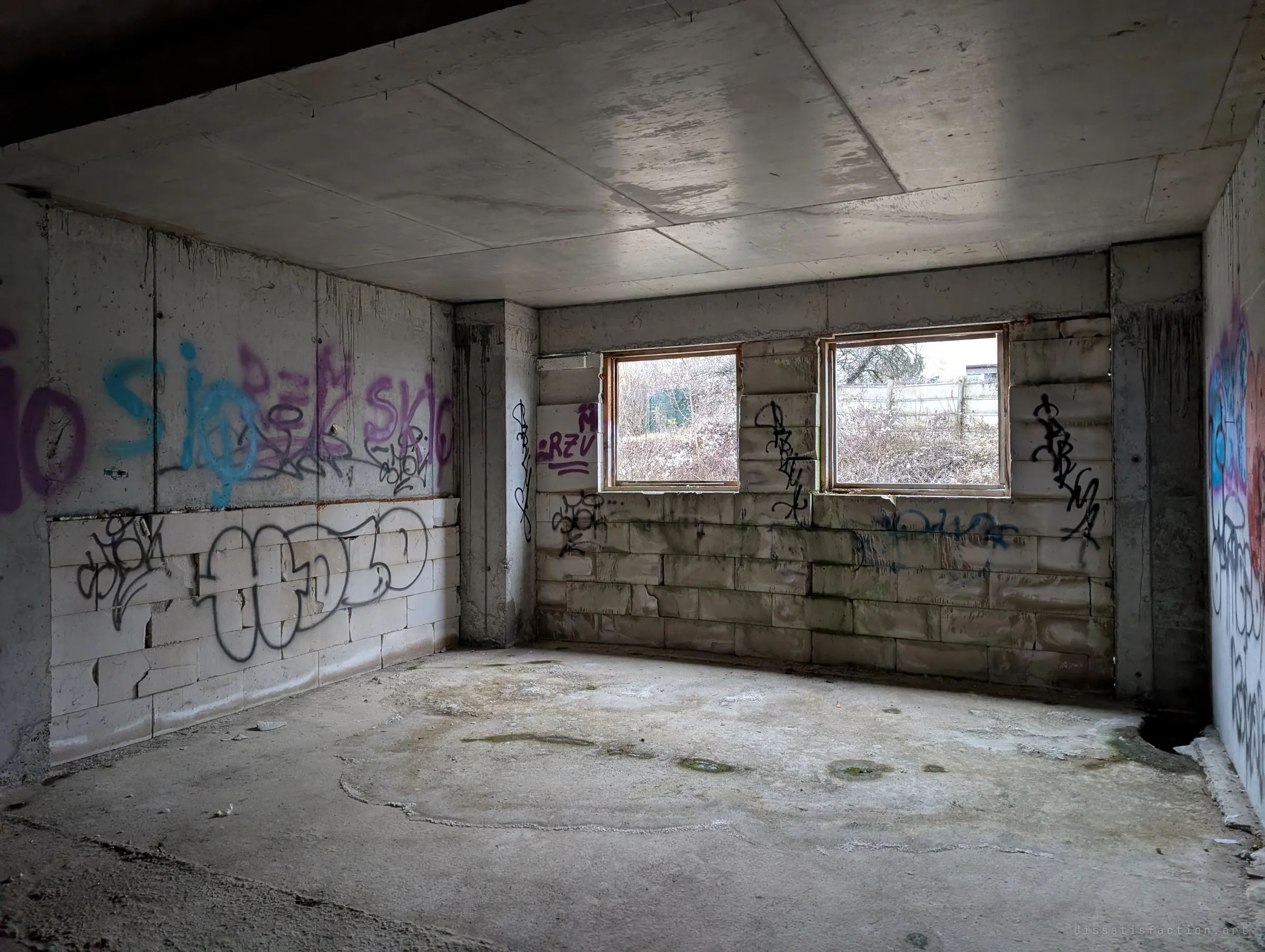 A graffiti-covered ground-floor room of block walls and cast concrete, two small windows looking out onto dry scrub.