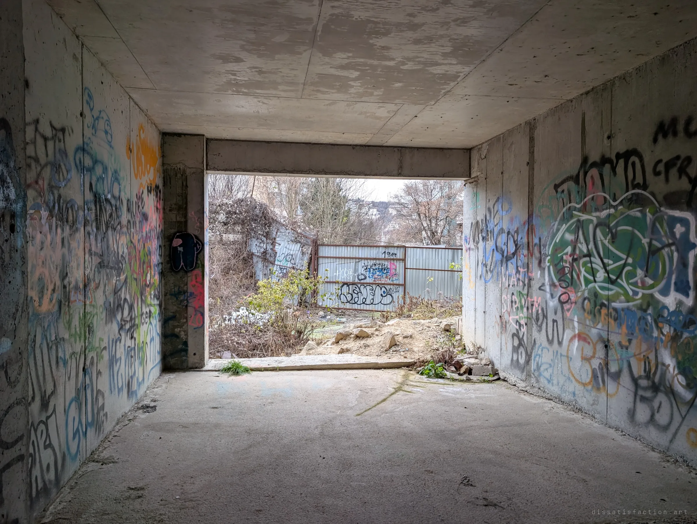 A tagged concrete passage opening onto a patched corrugated fence and overgrown rubble outside.