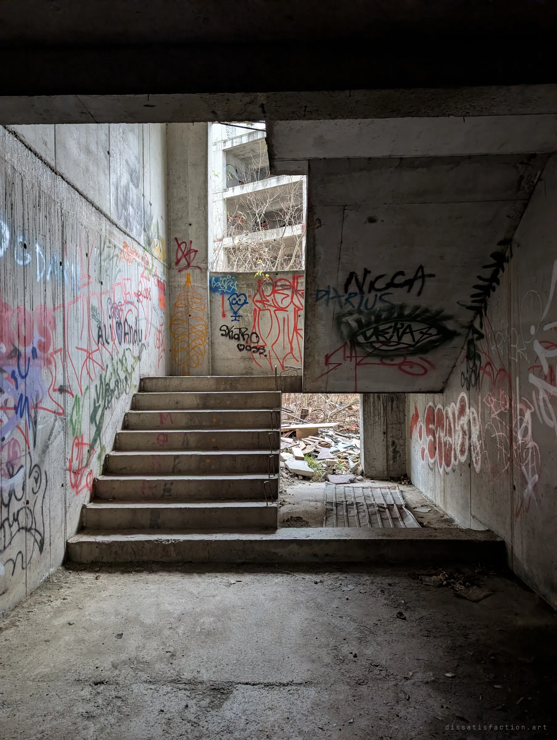 A graffiti-heavy stairwell climbing toward daylight, with the open frame of another unfinished block visible through the gap.