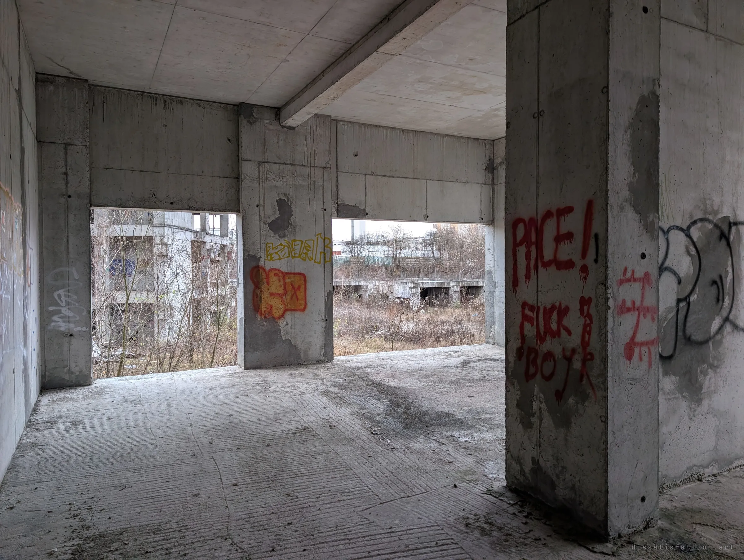 An open concrete floor with tagged pillars, its window gaps looking out on another half-built block and a low slab foundation in the weeds.