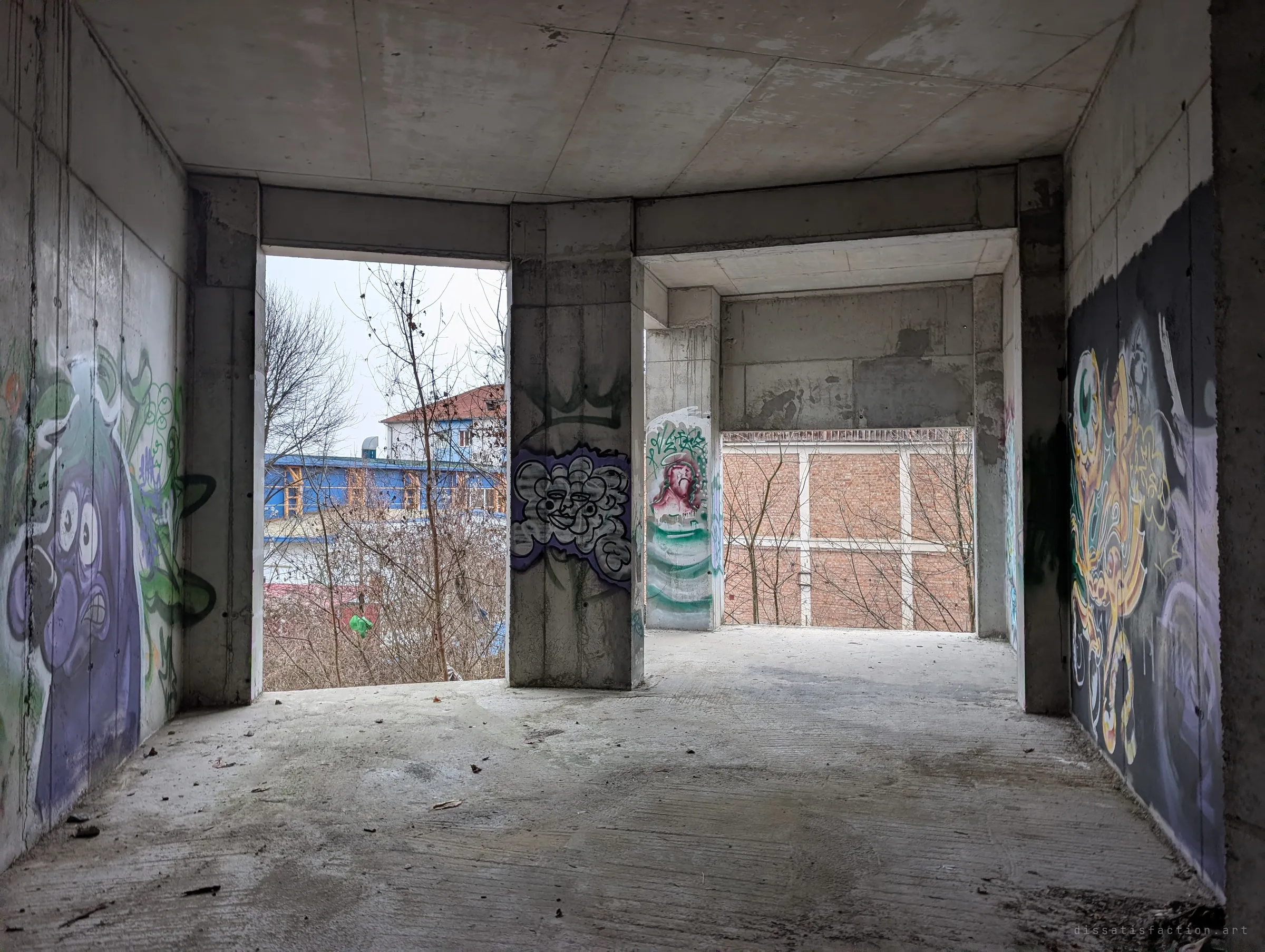 A bare room with large character murals on both side walls, its openings looking out on neighbouring buildings and a brick-infilled frame.