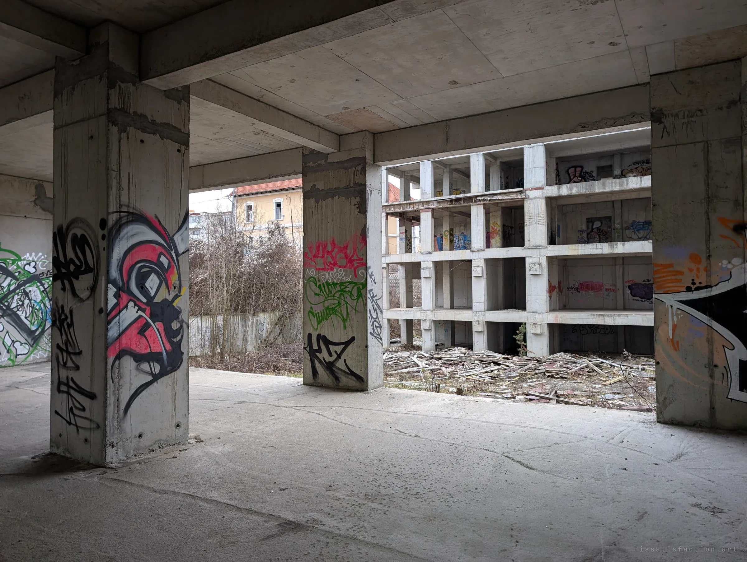 Painted pillars frame a view across to the skeletal grid of a neighbouring unfinished block, its ground strewn with broken timber.