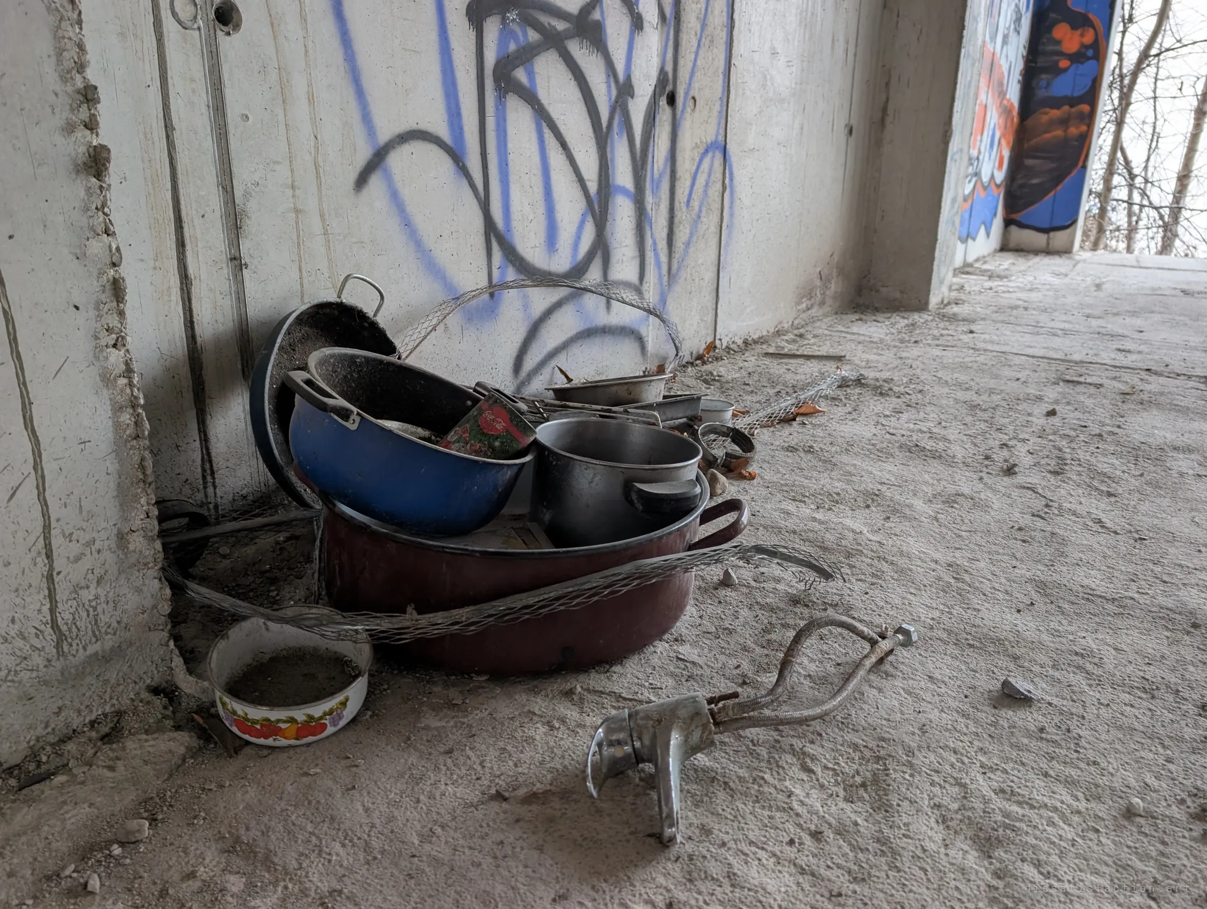 A pile of abandoned cookware and a detached tap left against a tagged wall, a quiet trace of someone once living here.