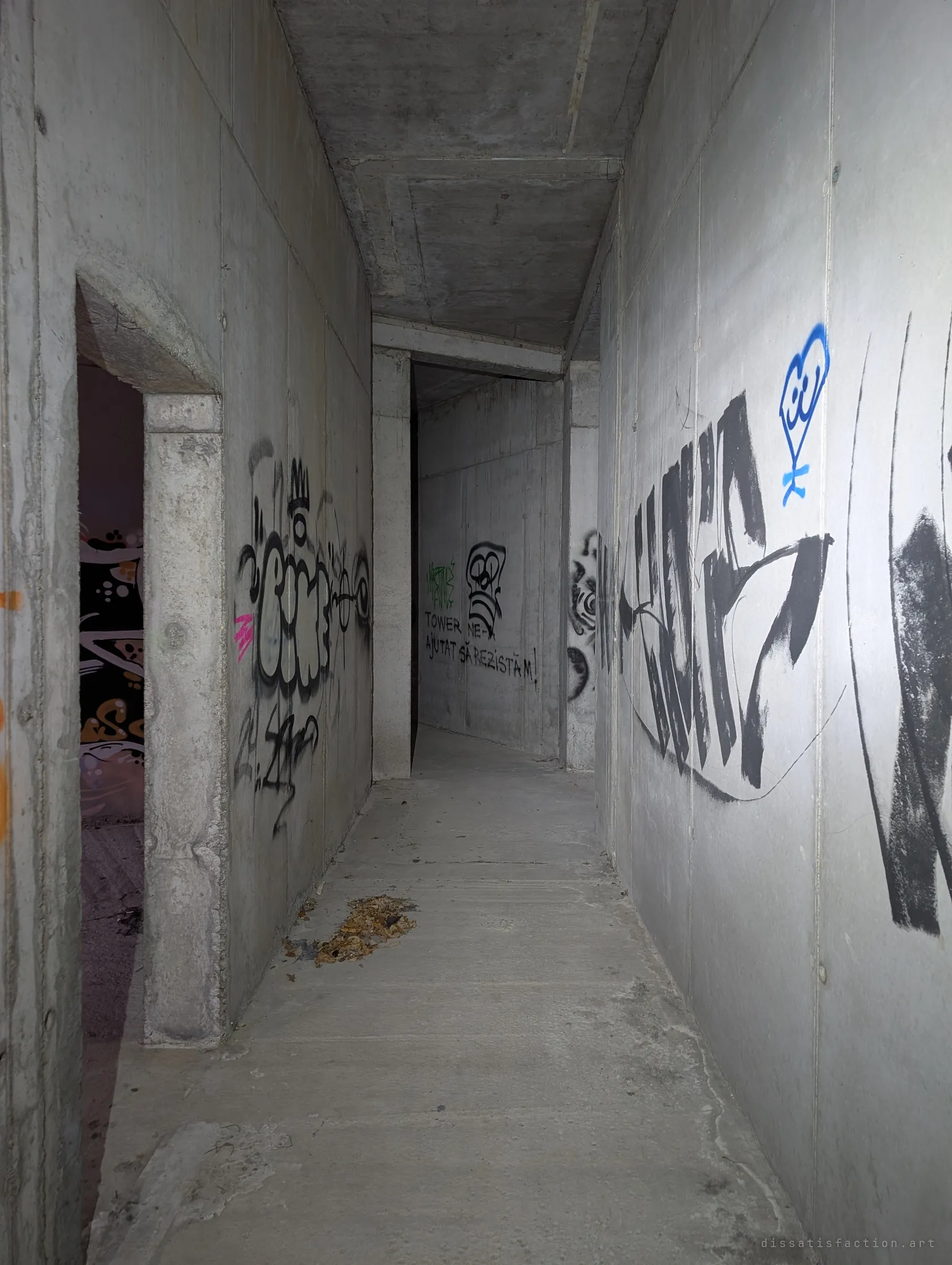 A narrow bare-concrete corridor lit by a torch, its walls covered in tags and a blue heart-faced doodle. The passages run deep into the unfinished shell.