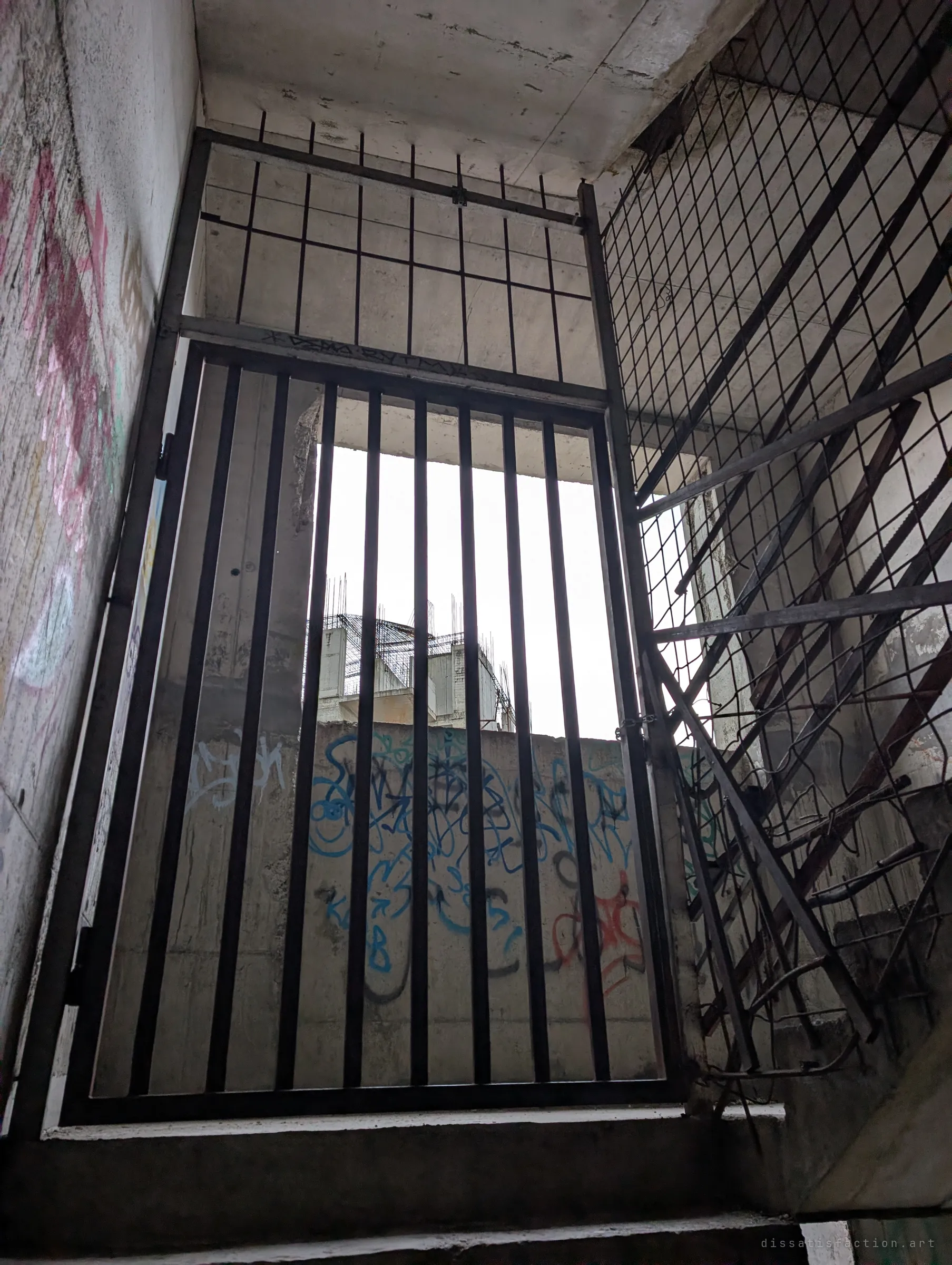 Looking up a stairwell to a barred steel gate and a torn mesh barrier blocking the way further up, with a rebar-crowned block visible through the bars. The route ends here.