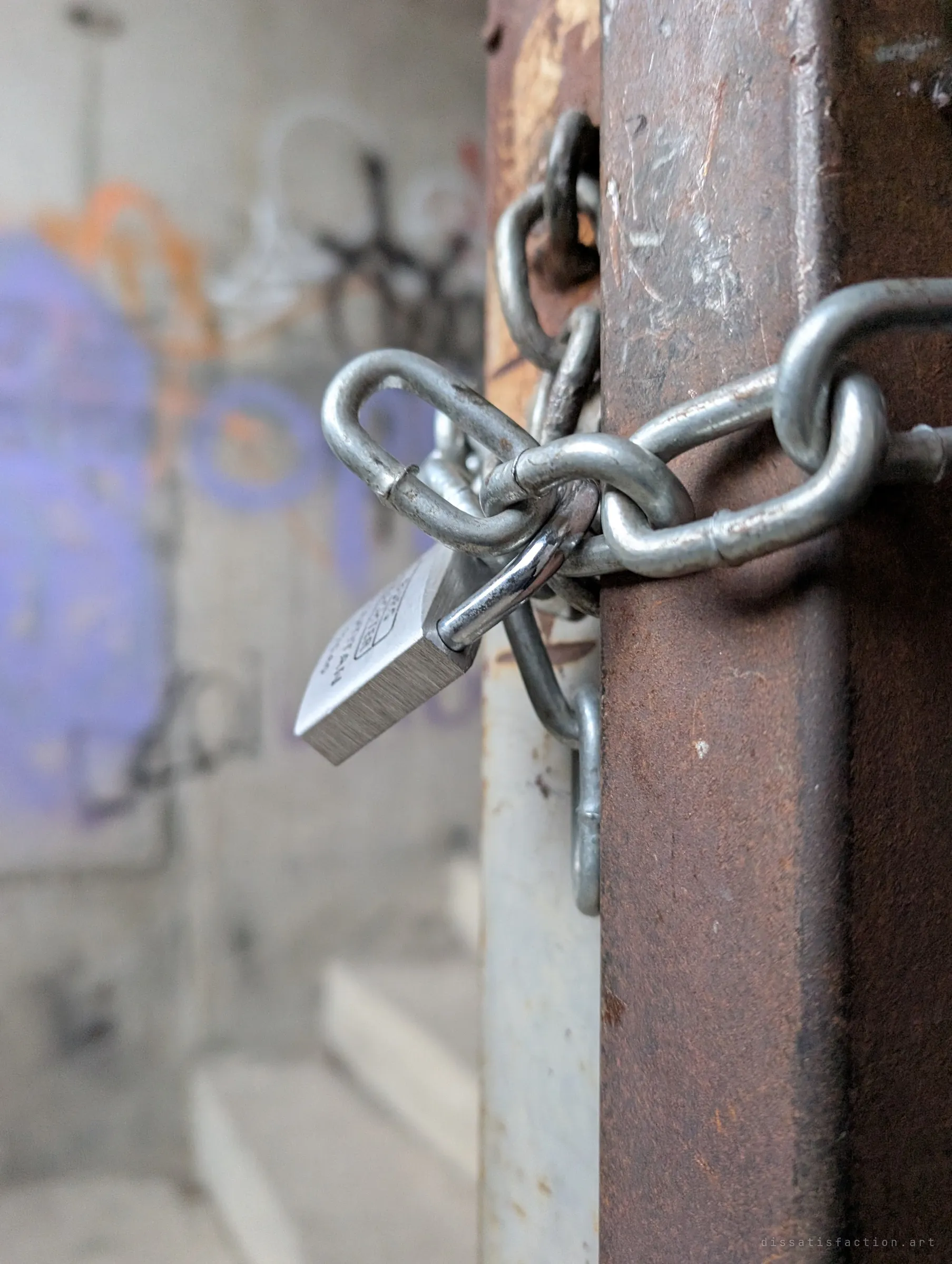 Close-up of a padlock and heavy chain wrapped around a rusted steel post, with blurred purple and orange graffiti behind. Someone still wants this place shut.