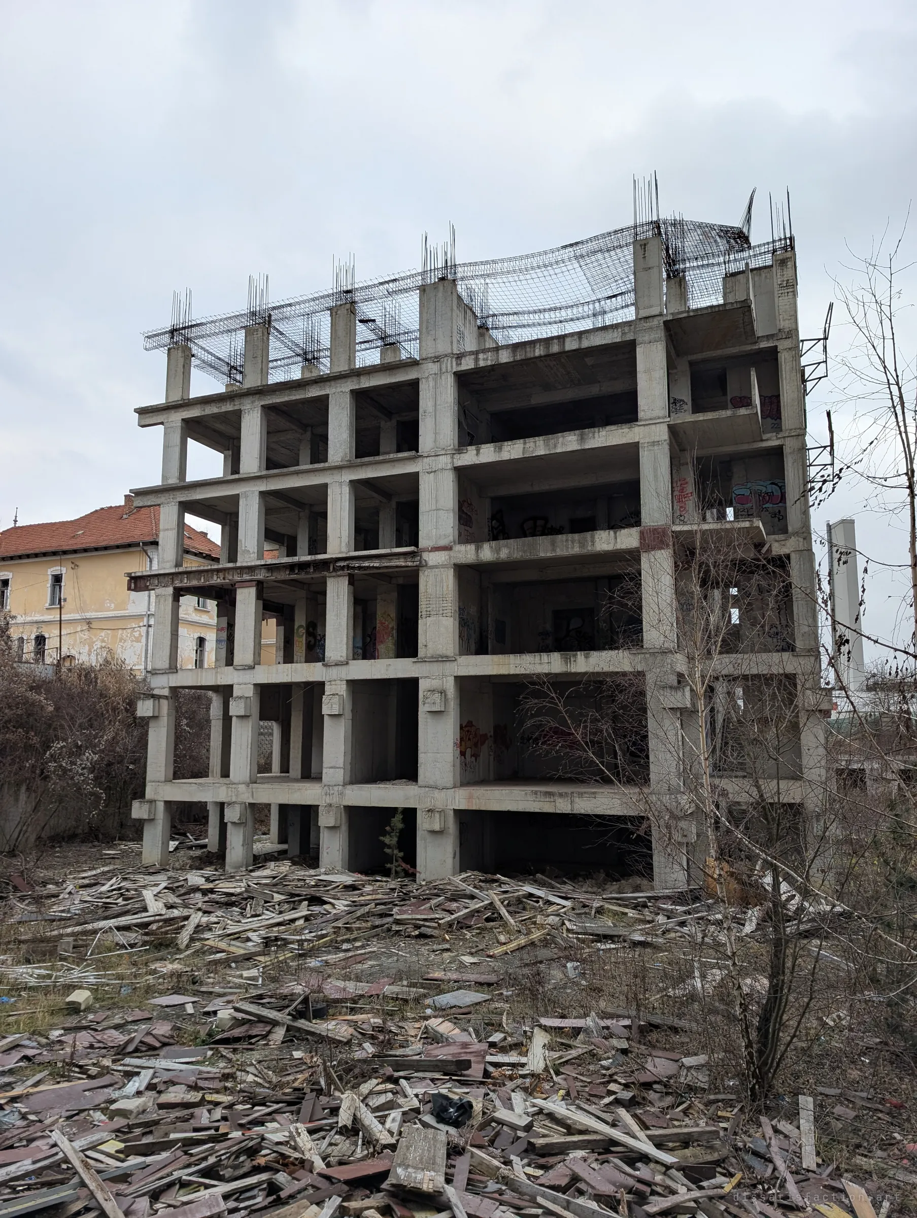 The raw concrete frame of an unfinished multi-storey block, rebar sprouting from its roof, standing over a field of scattered formwork planks. It shows the scale of the abandoned build.