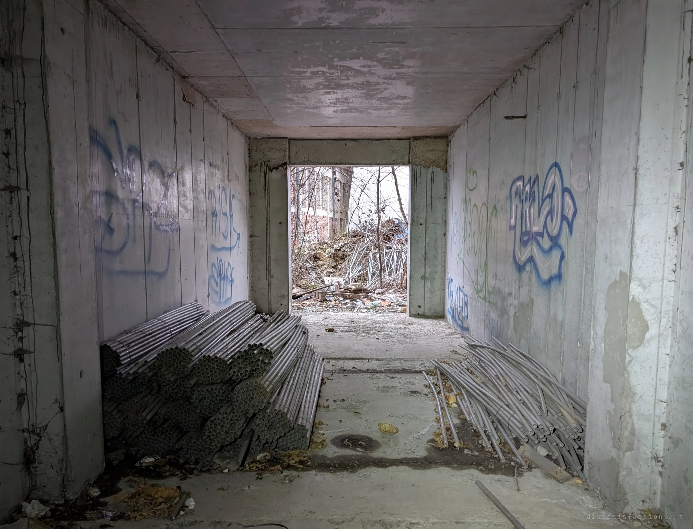 A concrete passage lined with bundles of abandoned conduit piping, opening onto rubble and bare trees outside. Materials left behind mid-build.