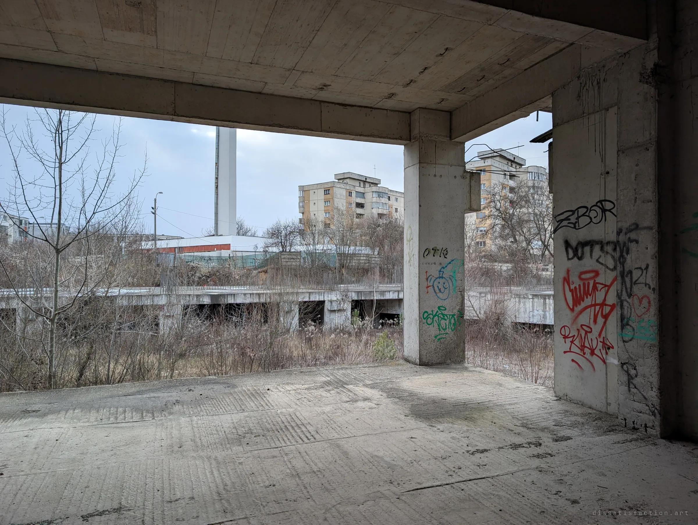 An open concrete floor looking out over overgrown lower foundations toward apartment blocks and a tall white stack. The unfinished site sits right inside a lived-in neighbourhood.
