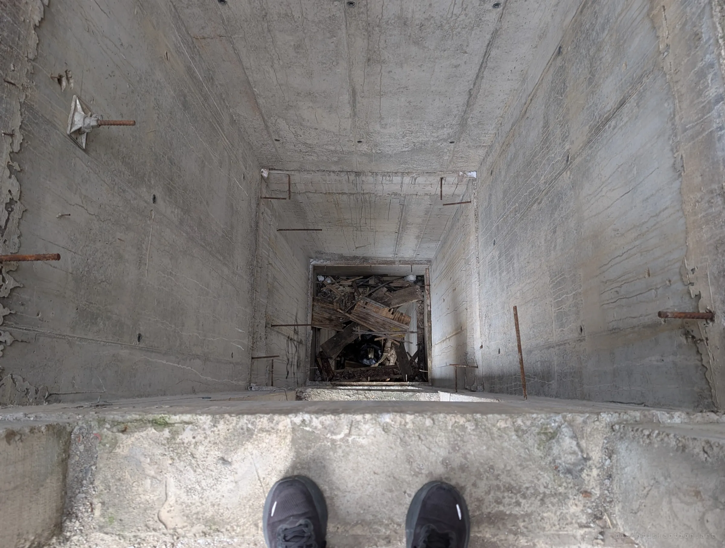 Looking straight down an open lift shaft from its edge, feet in frame, to a pile of broken planks at the bottom. There is no car and no railing.