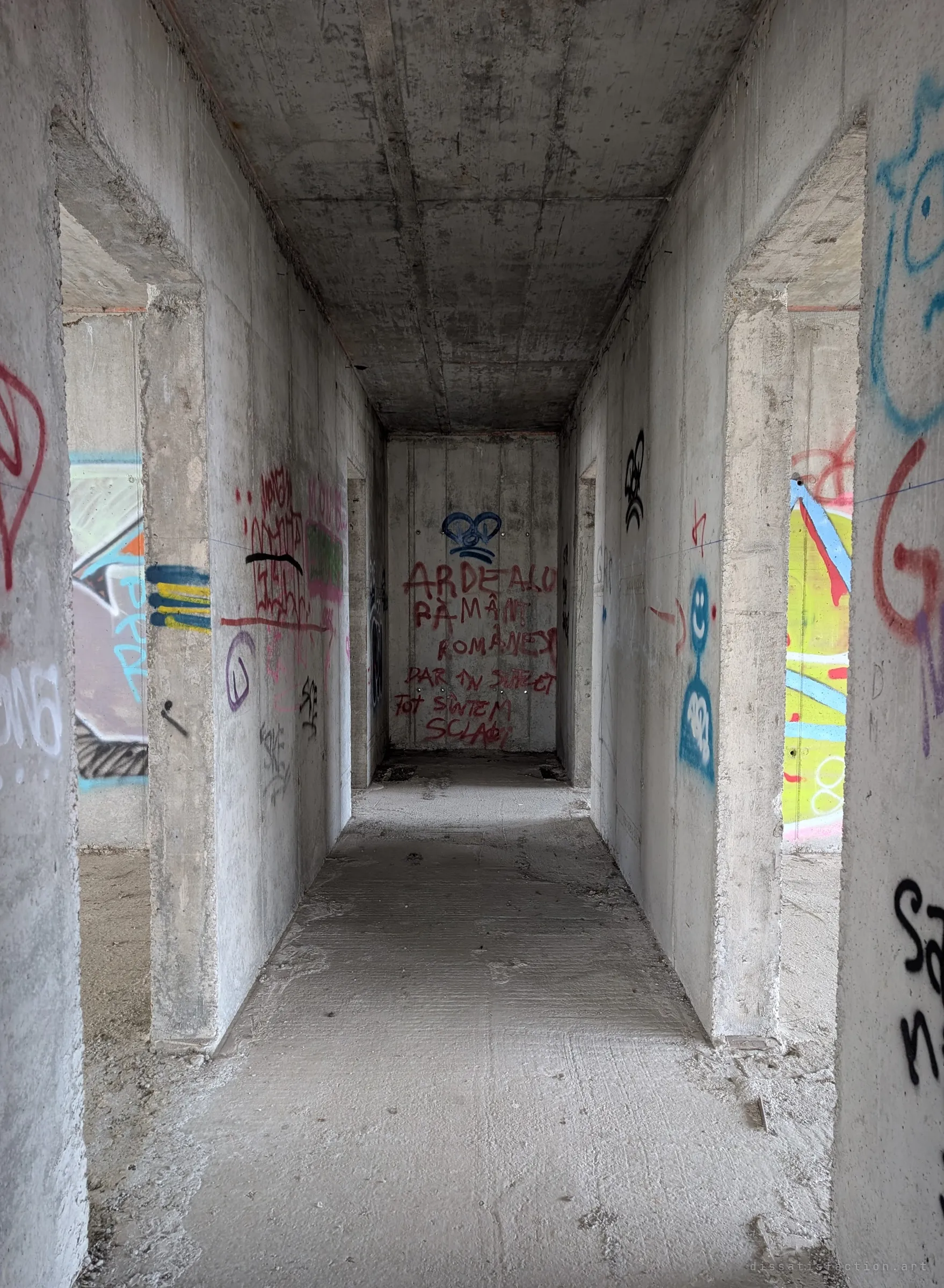 A bare concrete corridor runs between raw doorways to a back wall covered in red spray-painted words and a blue heart. The graffiti and grey shuttered concrete show how long the unfinished shell has stood open.