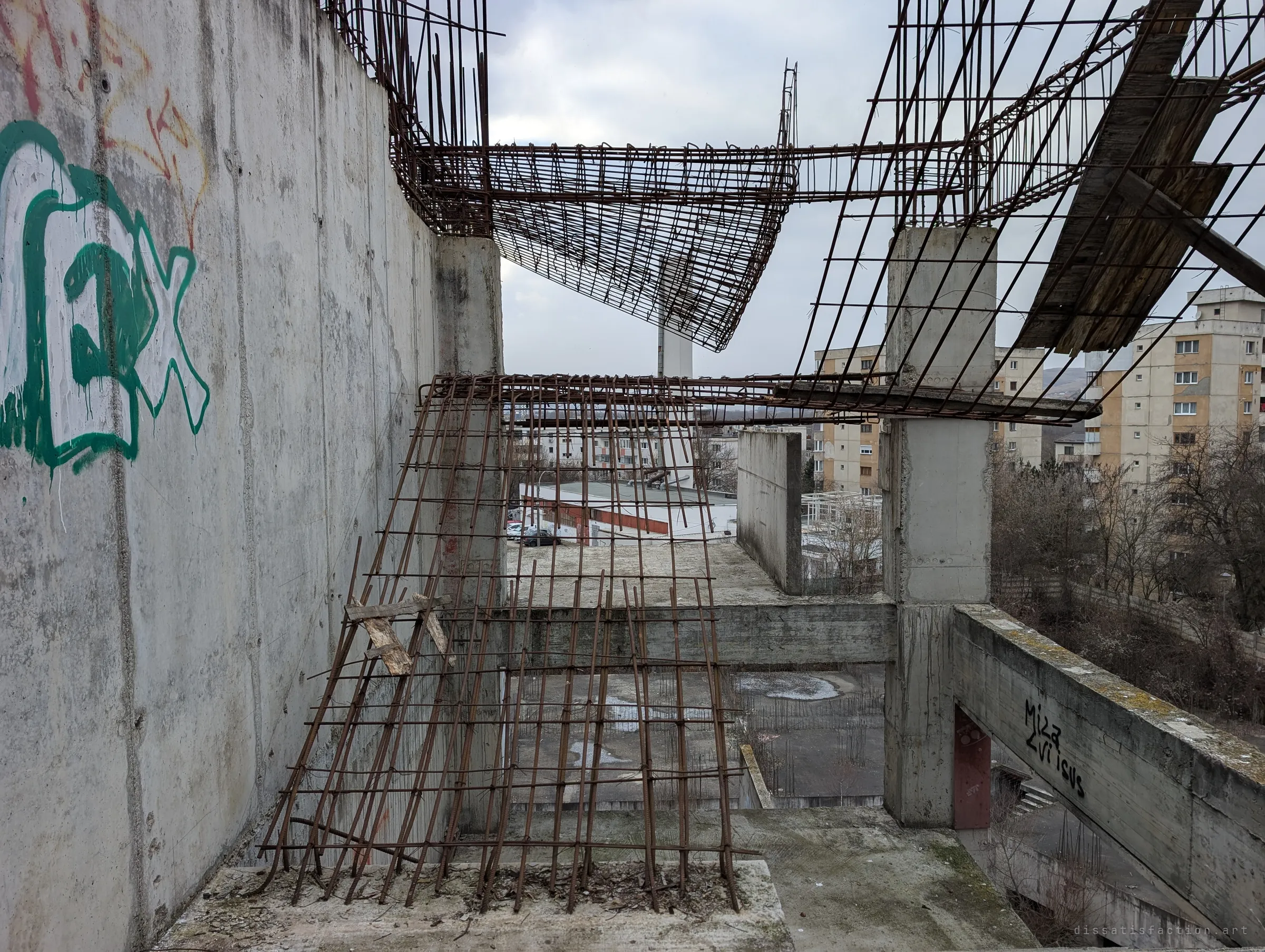 Bent rebar cages hang from the top of a raw concrete wall, sagging over an open floor like torn netting. Past them, ordinary apartment blocks carry on around the abandoned frame.