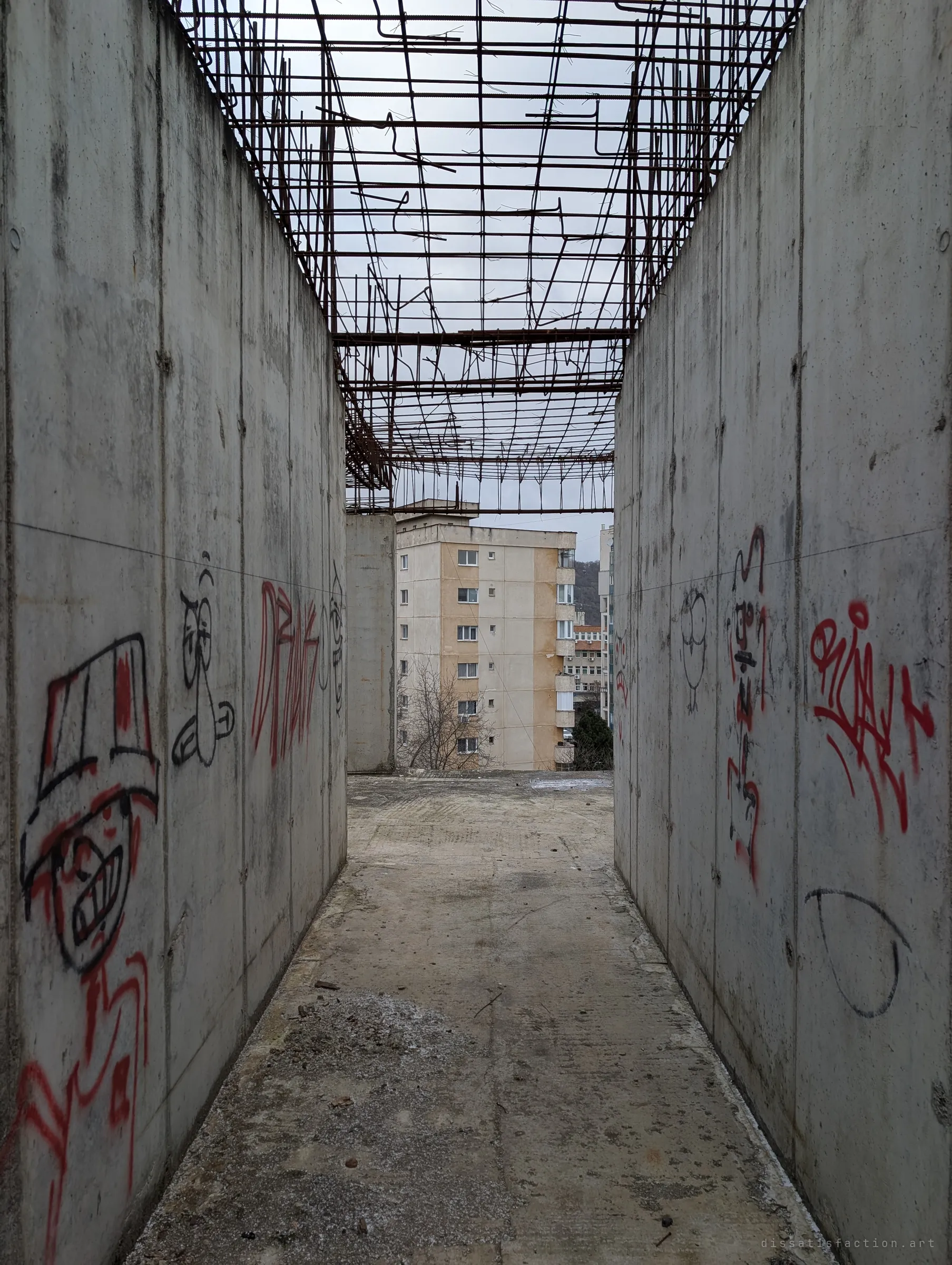 Two graffiti-covered concrete walls make a narrow roofless corridor under a loose grid of rusted rebar. At its end an occupied apartment block stands just beyond the ruin.