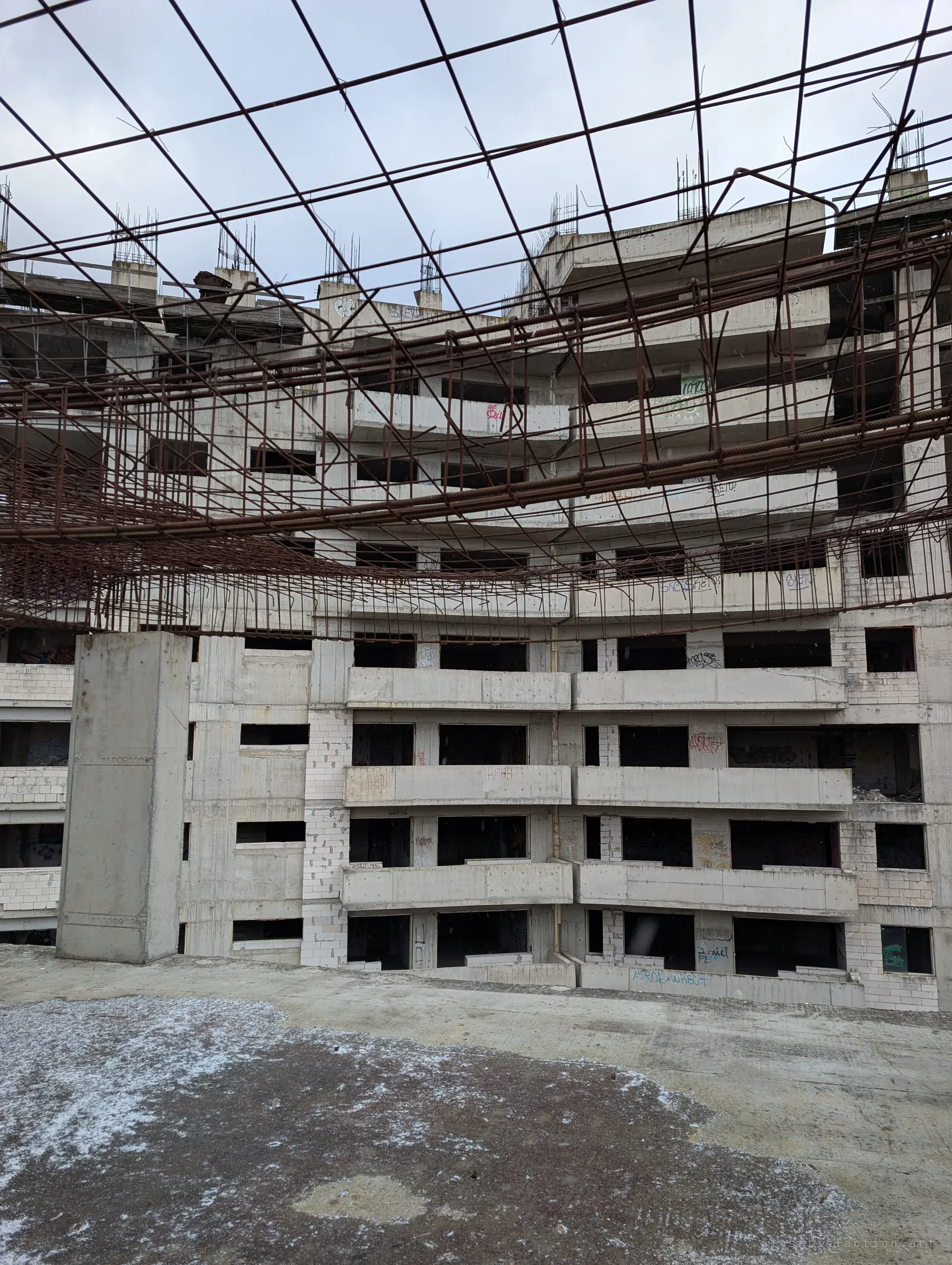 Seen through a sagging mesh of rusted rebar, a curved multi-storey concrete shell rises in tiers of empty balconies and window holes. Tags run along several floors of the never-finished block.