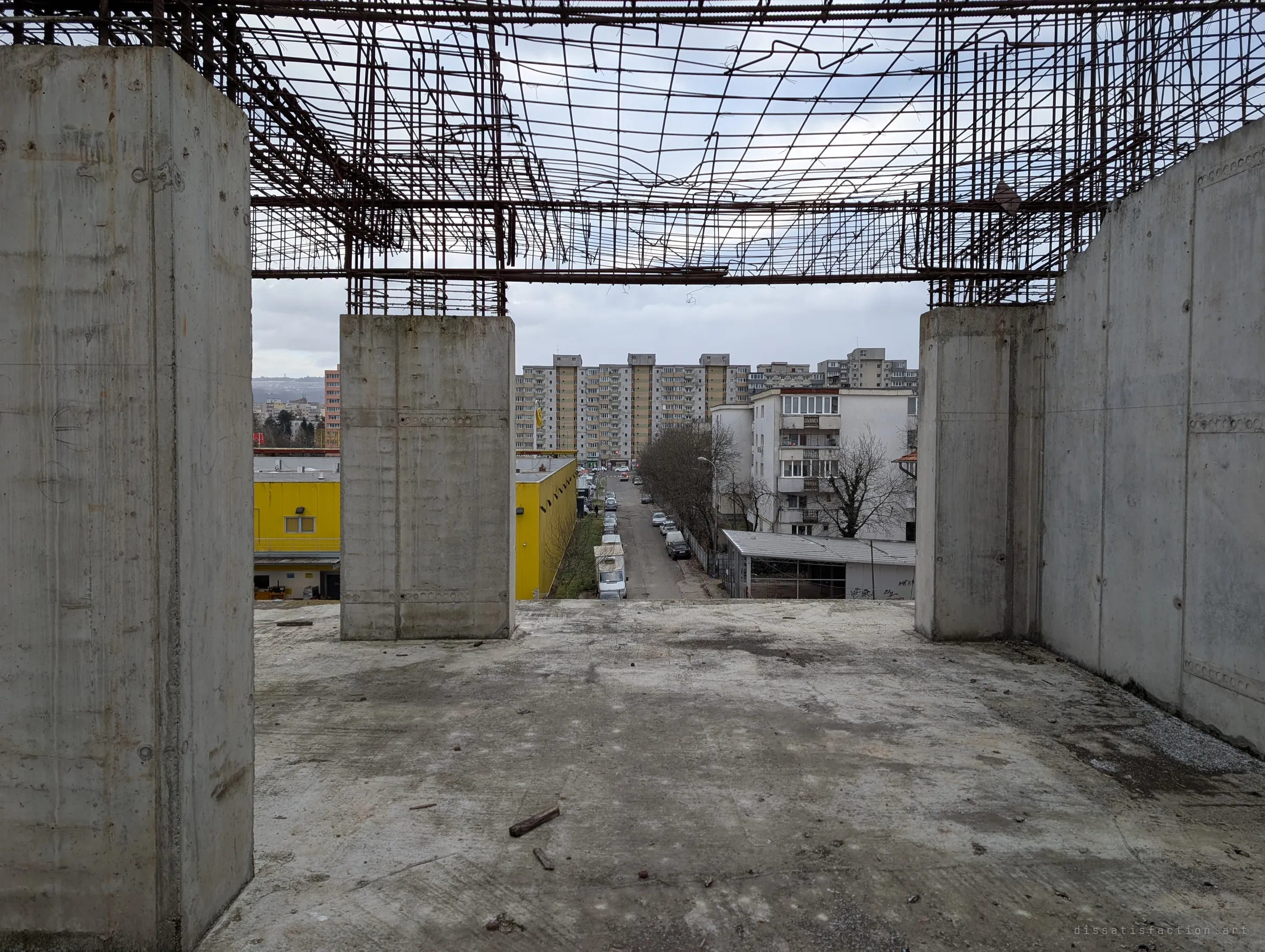 An open concrete deck, framed by two thick pillars and a ceiling of bare rebar, looks out over a street lined with parked cars and apartment towers. The half-built floor sits right above everyday city life.