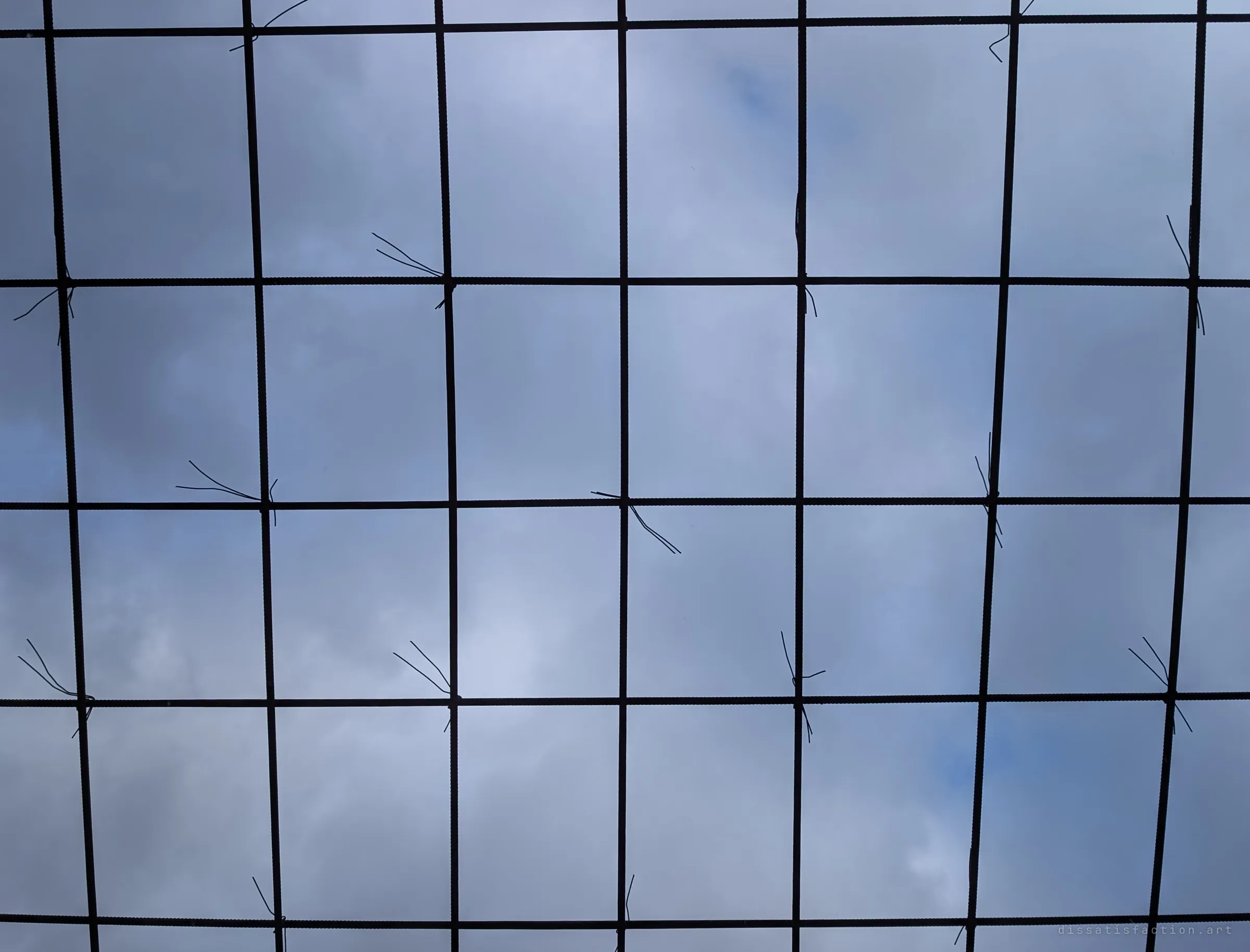 Looking straight up through a square grid of tied rebar at an overcast sky. The roof was never poured.