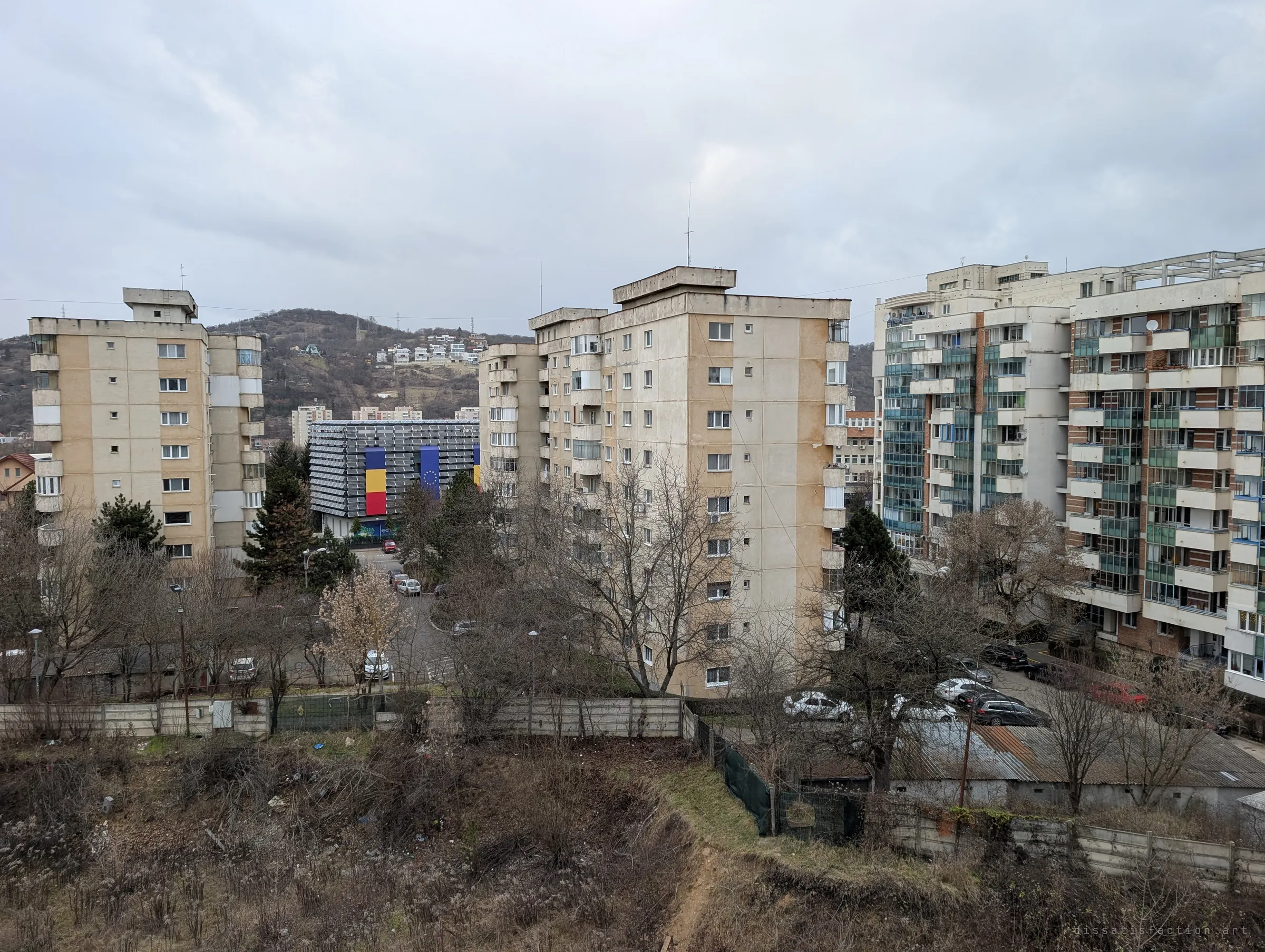 Tall apartment blocks stand behind a fenced, overgrown lot below the ruin, with a wooded hill beyond. The view shows how the abandoned site is wedged into a lived-in neighbourhood.