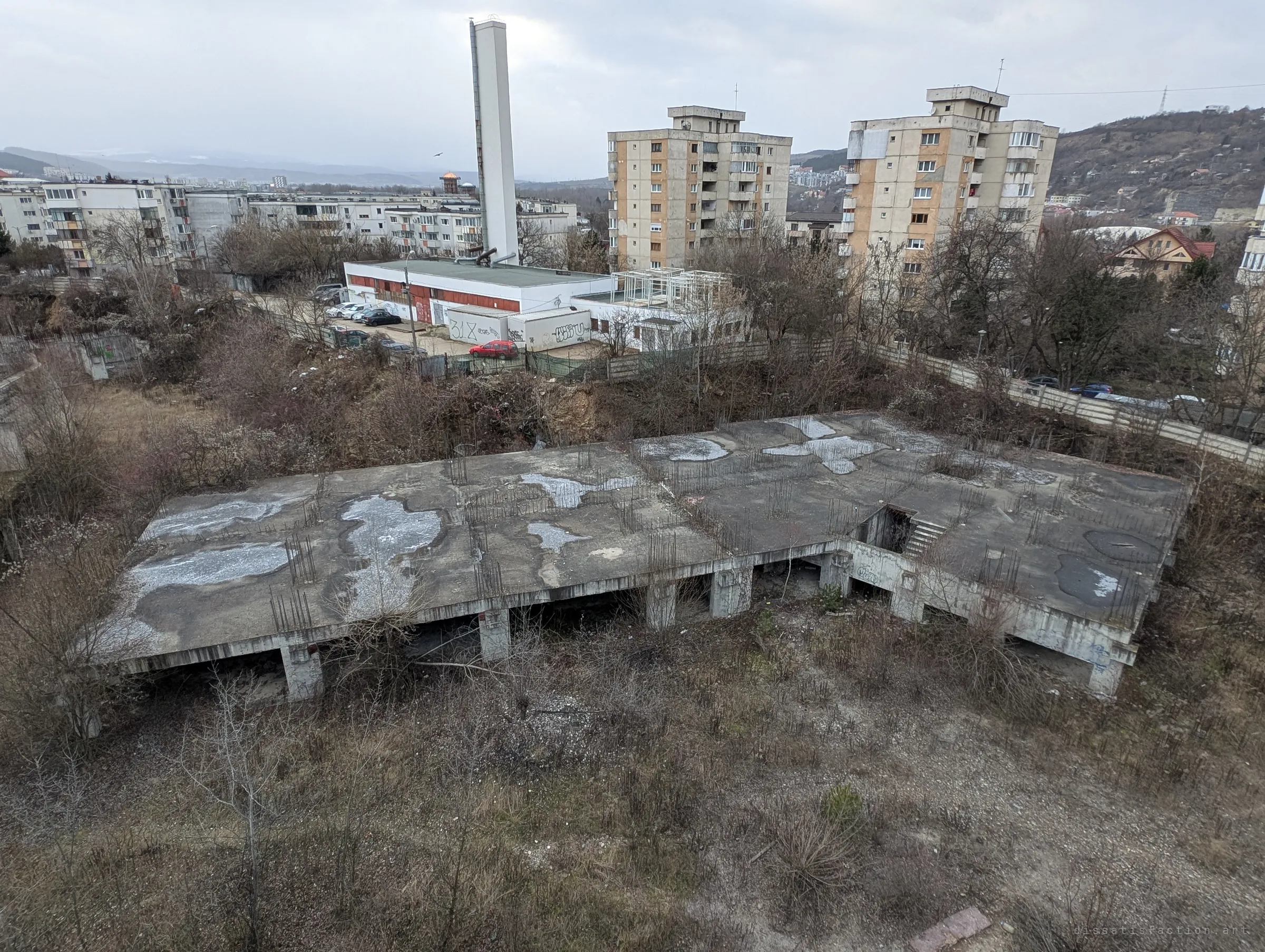 From above, a long flat concrete slab sits on stubby pillars in a field of dry scrub, with puddles and rebar stubs on its roof. It is the lowest block of the complex, abandoned at its first floor.