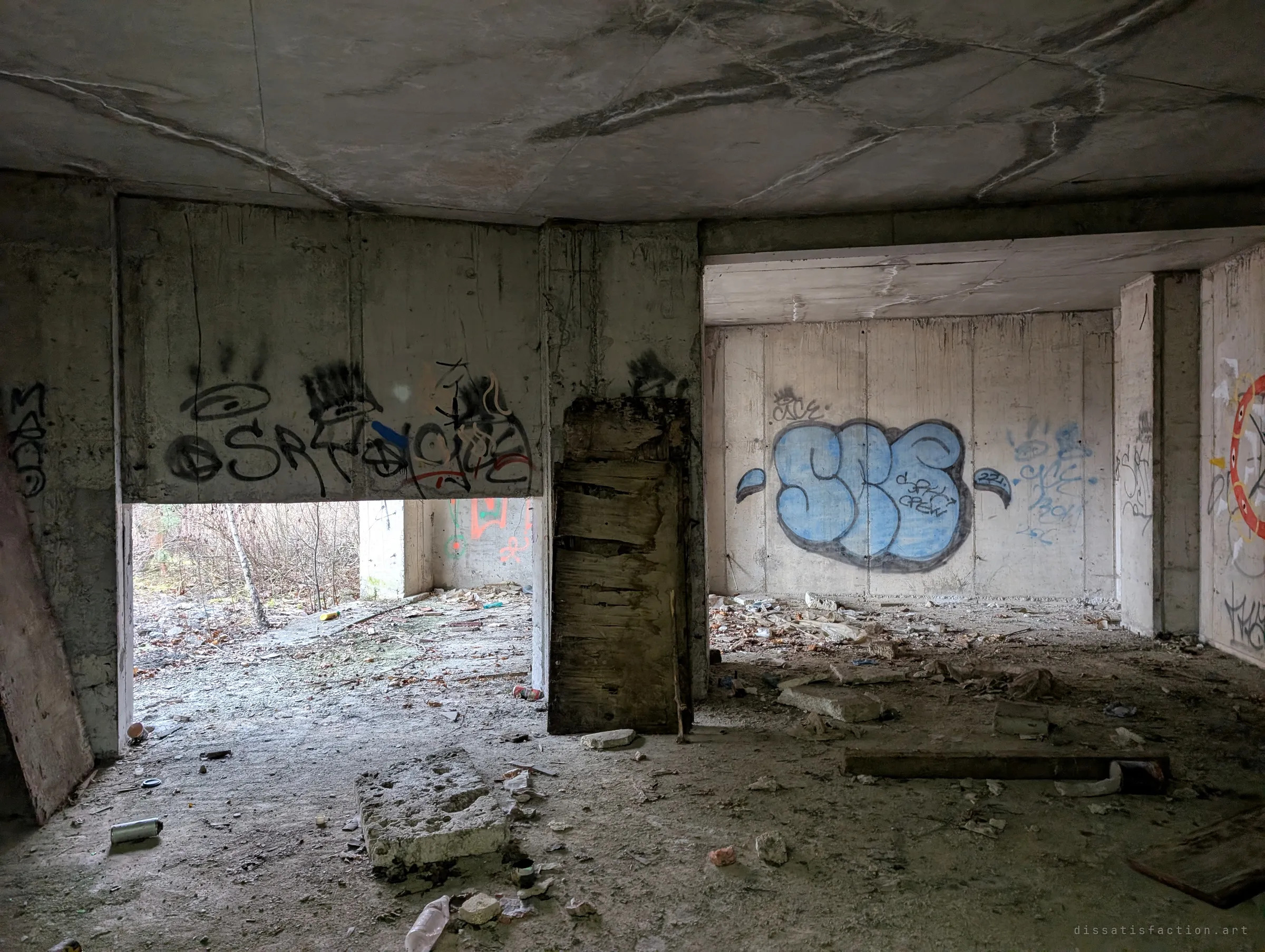 A dim ground-floor room littered with rubble and spray cans, its walls covered in tags and a large blue throw-up. A rotting board leans against a pillar in front of an opening onto bare trees.
