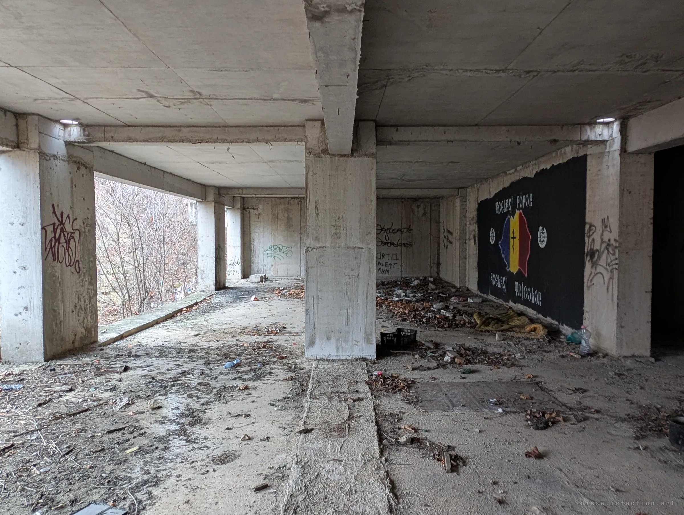 An open ground-floor hall of raw pillars and beams, strewn with leaves and rubbish, with a black mural of a painted tricolour map on the back wall. An old blanket and bottles near the mural hint that people shelter here.