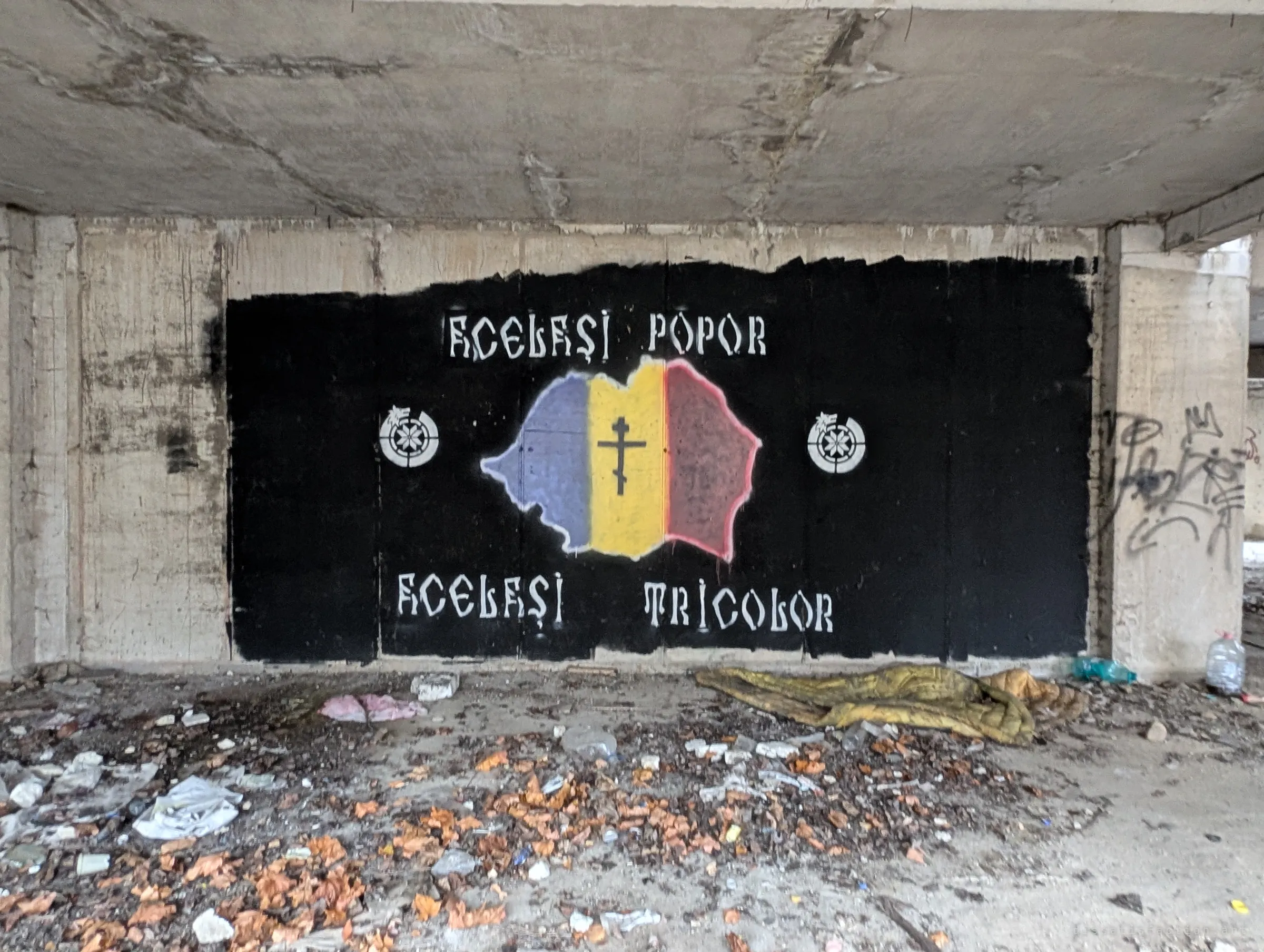 A black-painted wall carries a nationalist mural of a tricolour country outline with an Orthodox cross and slogans, above a floor littered with leaves, rubbish and an abandoned sleeping bag.