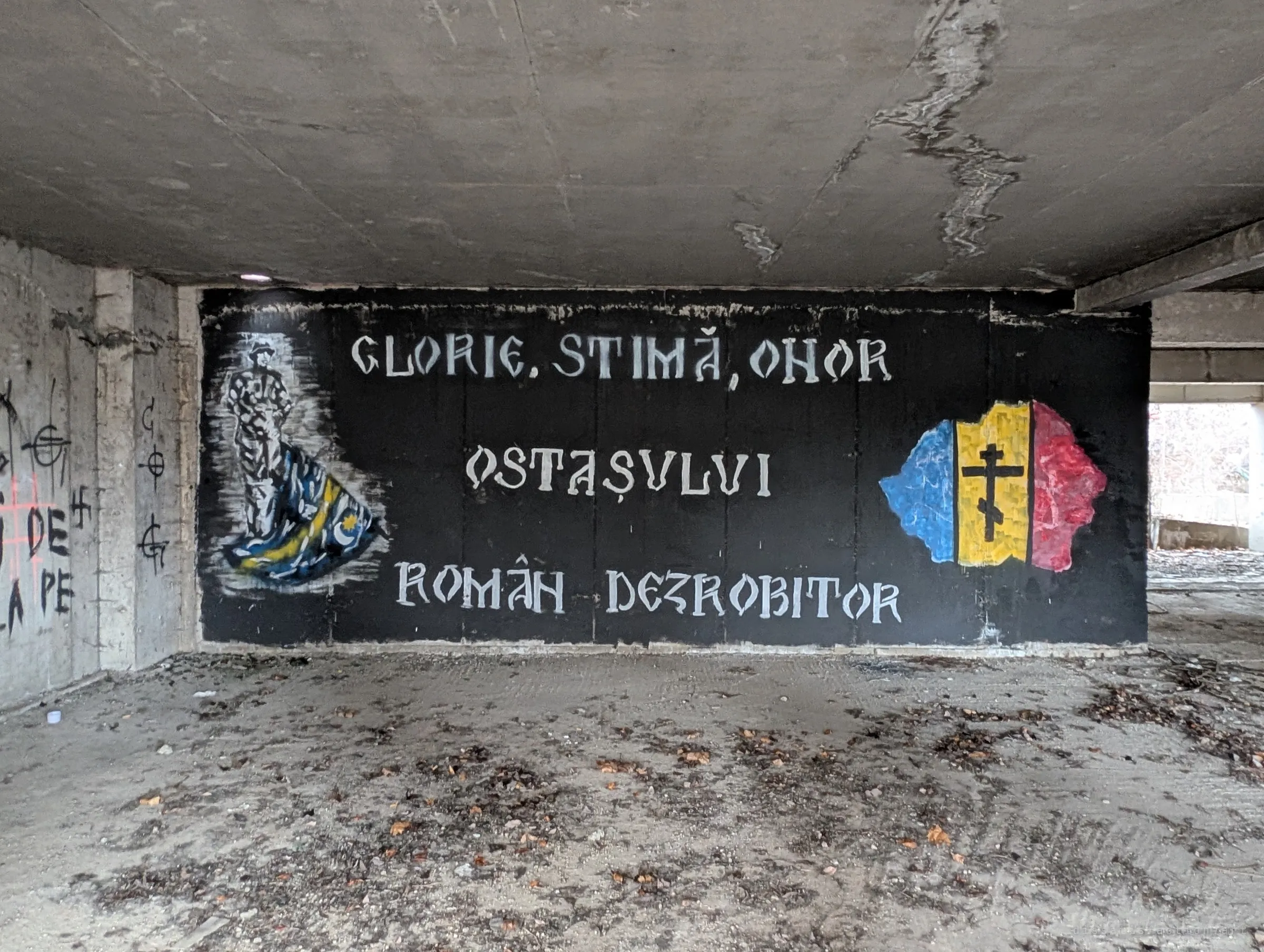 Another black mural under the concrete slab honours a soldier with painted slogans and a flag-coloured map bearing a cross. The bare floor and open bays behind show the building never got past its frame.
