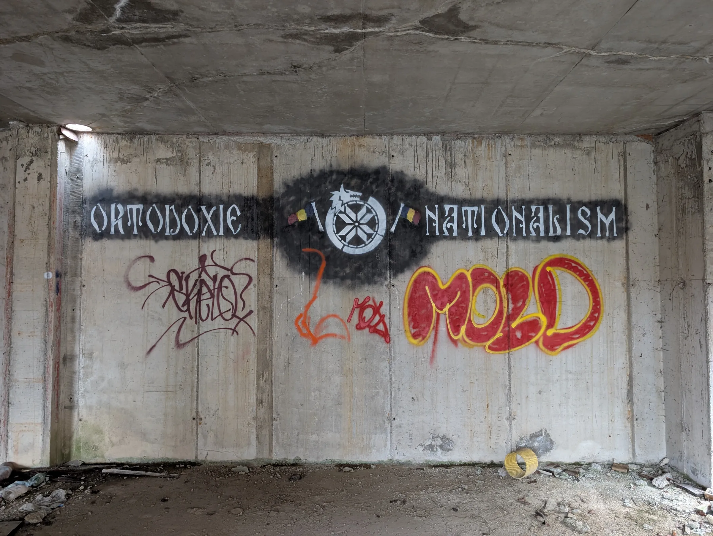 A stencilled banner of religious and nationalist slogans runs across a concrete wall, overlapped by colourful graffiti tags. A light well lets a pale circle of daylight in at the corner.