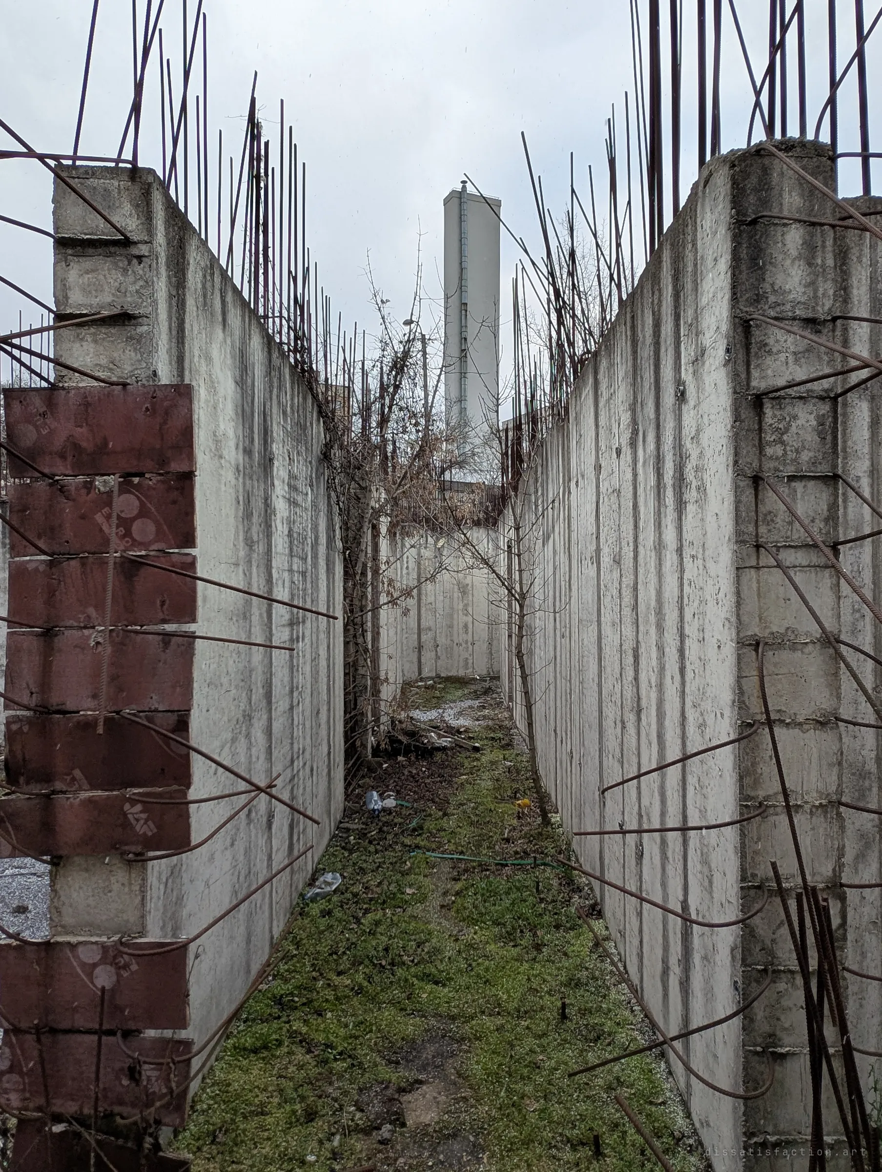 A narrow mossy corridor runs between two concrete walls bristling with bent rebar, leading the eye to a tall pale tower in the distance.