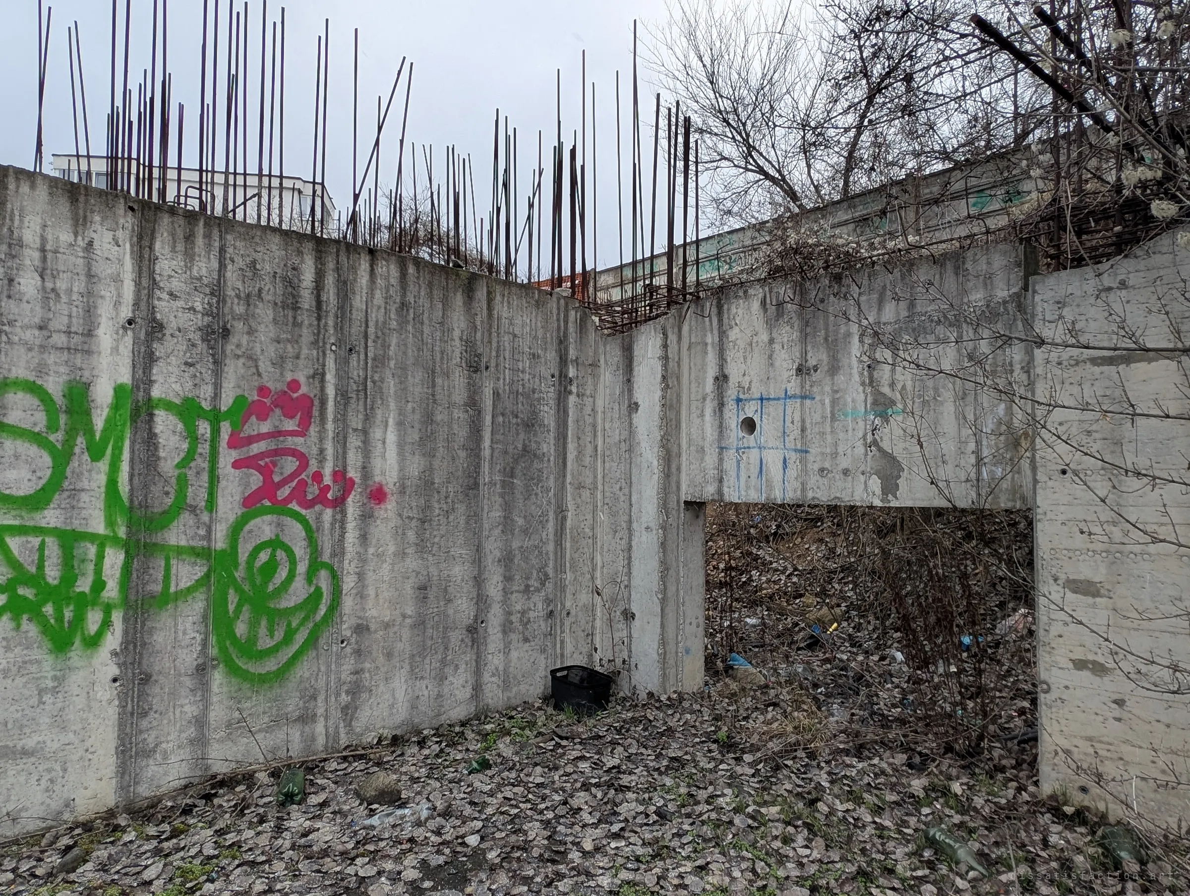 Graffiti-sprayed retaining walls with rebar along the top wrap around a leaf-covered floor, and an opening leads into a rubbish-filled void under the slope.