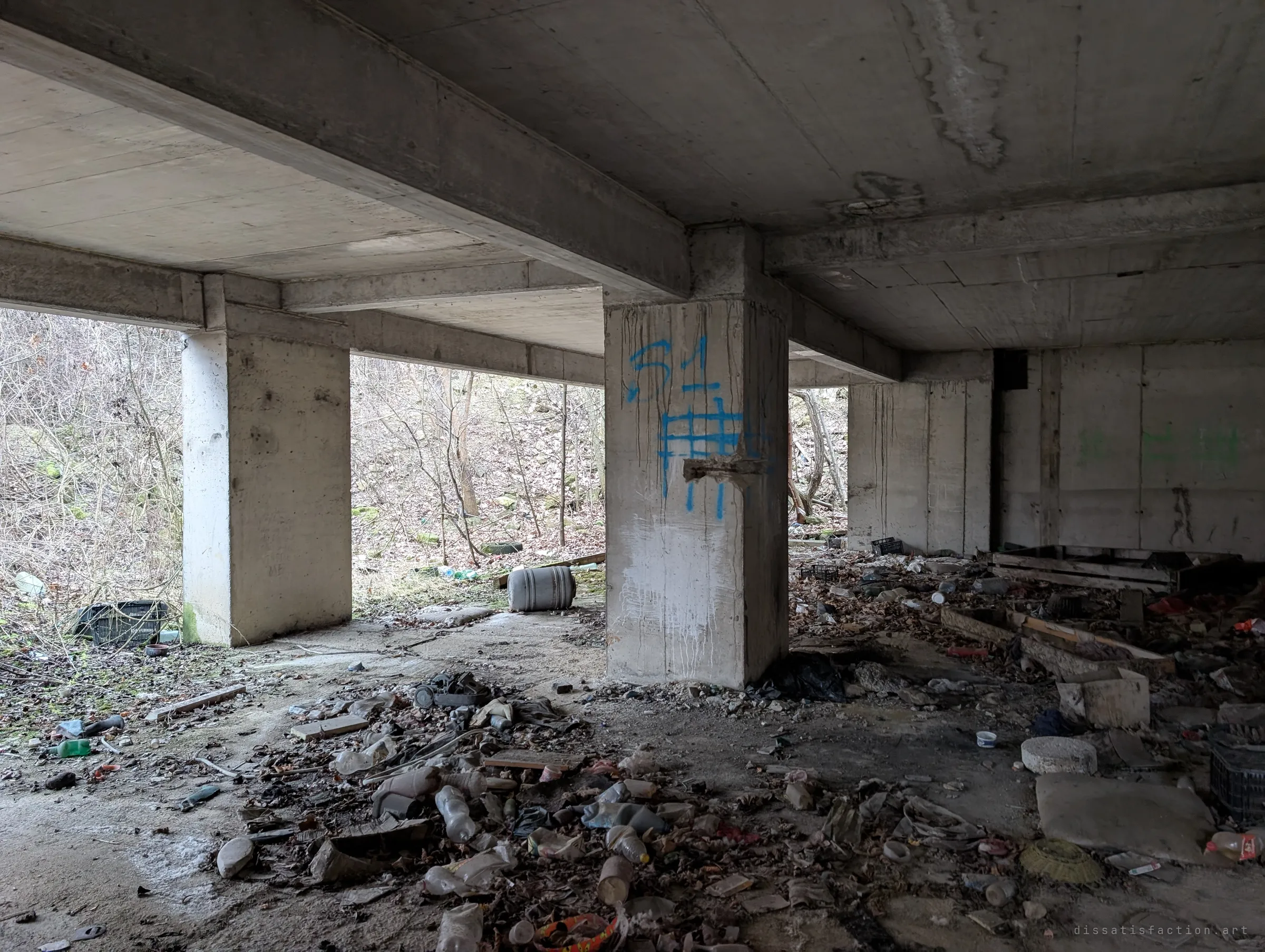 Under the open concrete frame, columns and beams stand over a floor strewn with bottles and rubbish, with bare woodland just outside. Signs of people camping here.