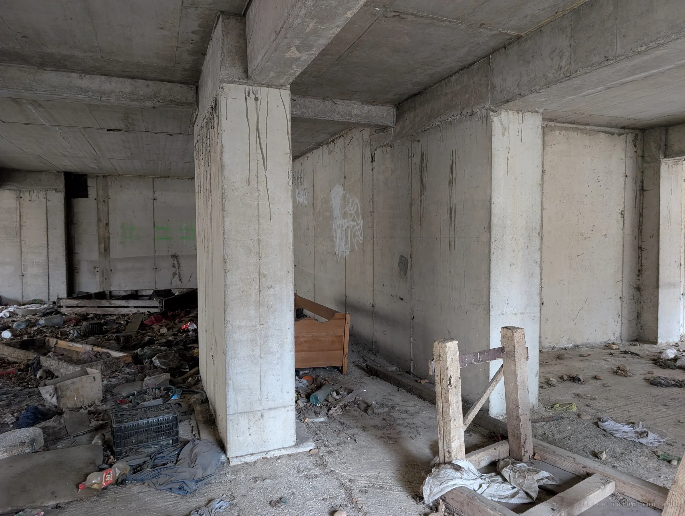 Inside the ground floor, a concrete wall and columns split the space; an old wooden bed frame, crates and debris show people once sheltered here.