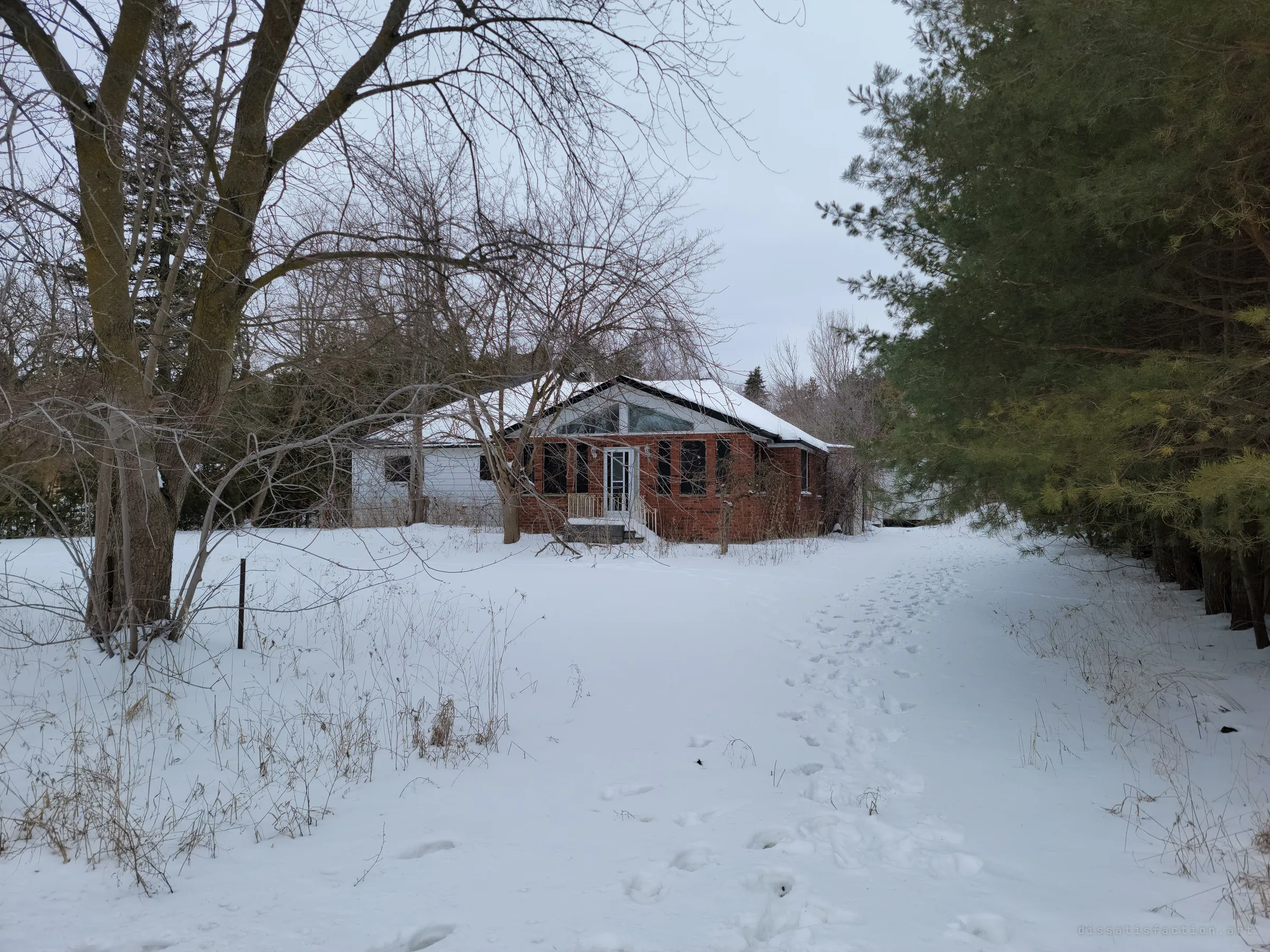 A small brick-fronted house with a glassed gable sits behind bare trees at the end of a snow-covered lane, footprints leading up to it. It sets the scene: a rural home left to winter.