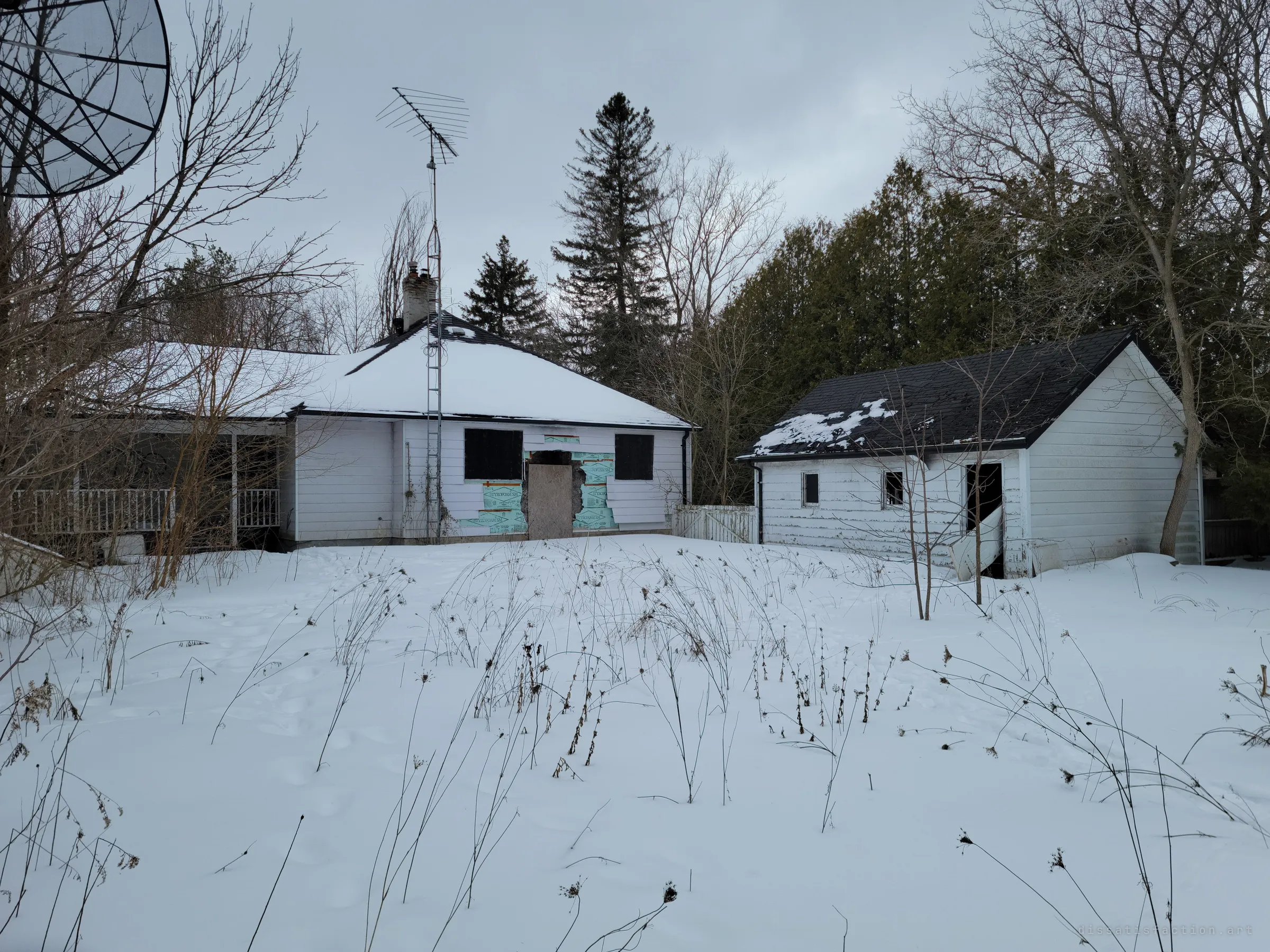 The back of the white-sided house, its rear door boarded and wrapped in torn house wrap, beside a detached garage with a broken door, all behind a snowed-over yard of dead stalks and an old antenna mast.