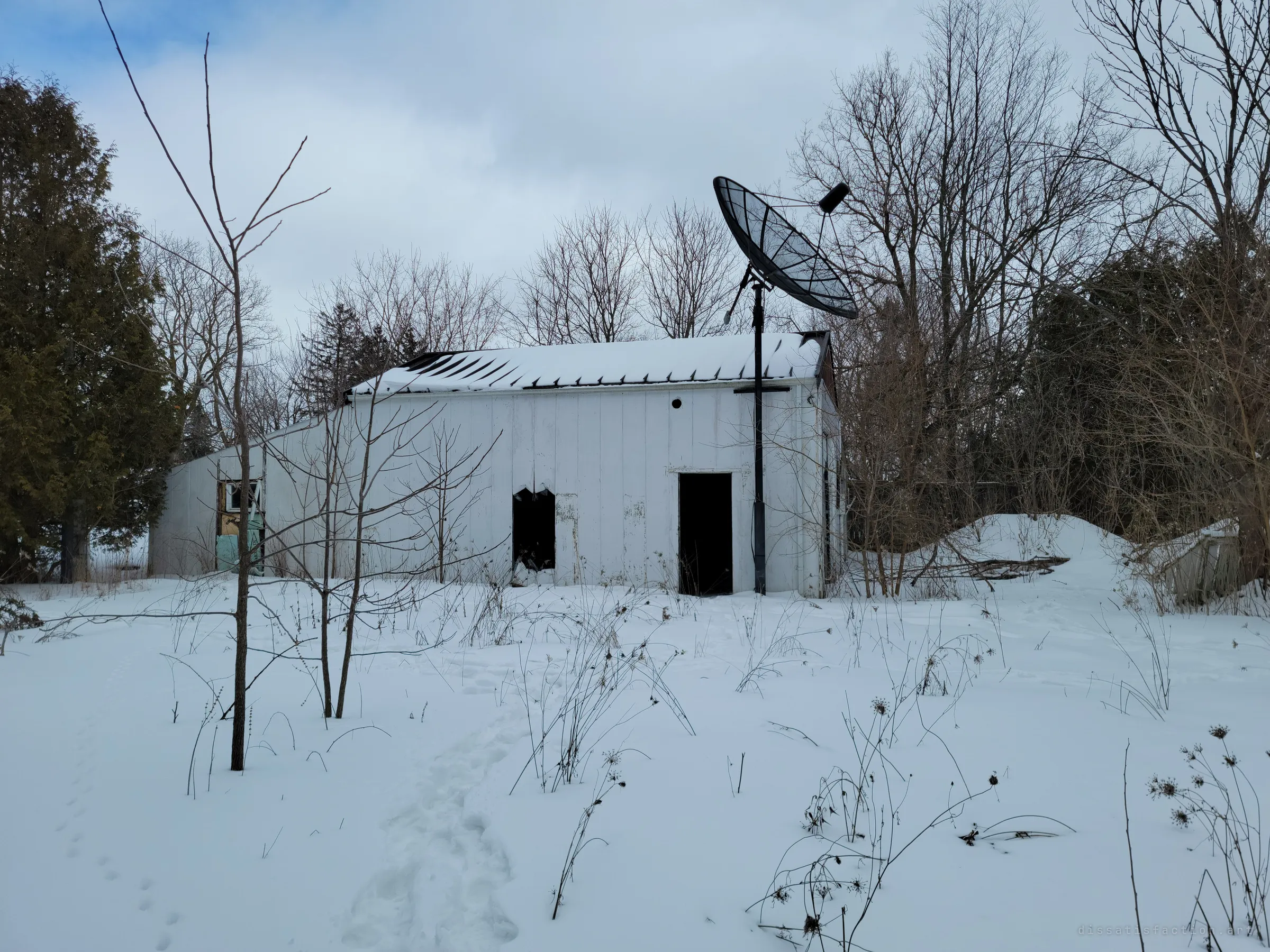 A long white outbuilding with a sagging, peeled-back roof stands in deep snow, an old mesh satellite dish tilted on a pole in front of it. Footprints lead to its dark open doorway.