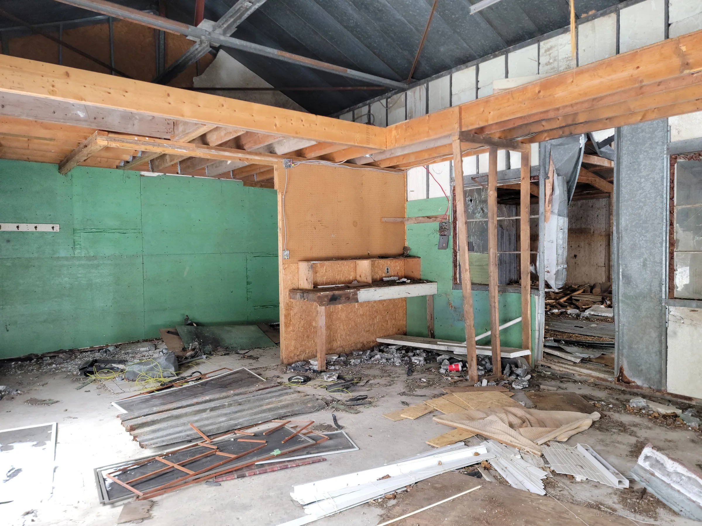 A gutted workshop with green-painted walls, a pegboard workbench and exposed joists, the concrete floor littered with window screens, blinds and rubble.