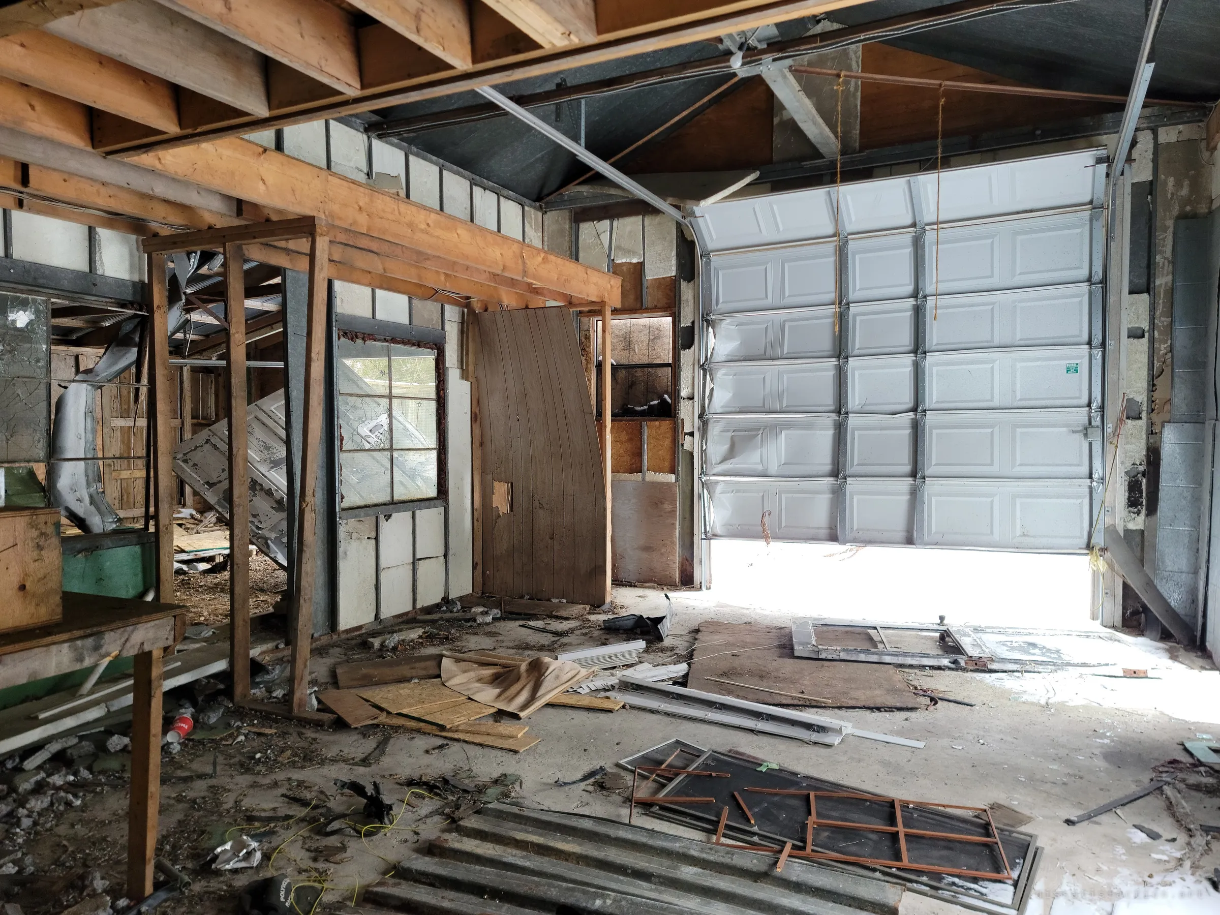 The same workshop looking toward a dented overhead garage door, jammed half open so daylight floods across the debris-strewn floor and broken framing.