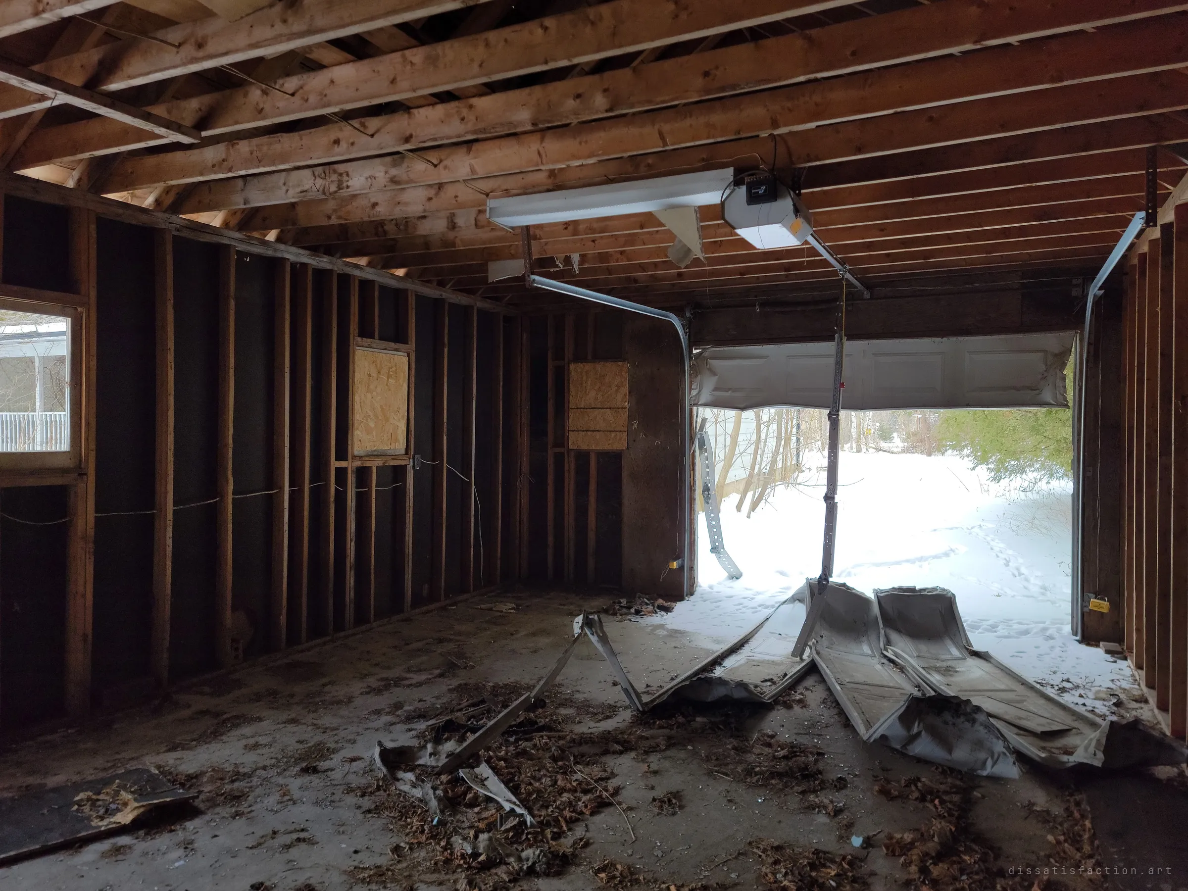 Inside the stripped detached garage, open studs and boarded windows frame a garage door torn from its track, its crumpled panels spilling out onto the snow.