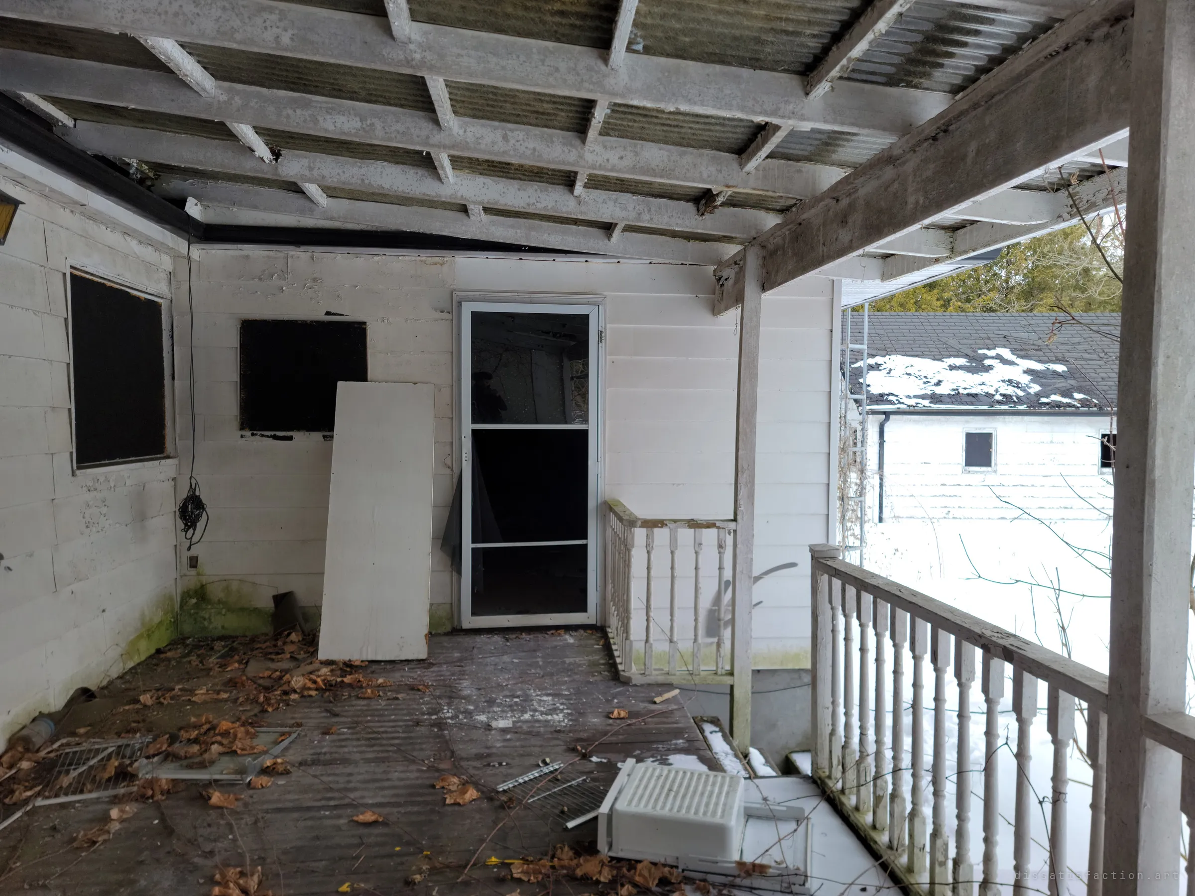 A covered back porch with peeling white siding, a propped-up board and a broken screen door, leaves and a discarded air conditioner on the boards. Snow lies on the roof of an outbuilding beyond the railing.