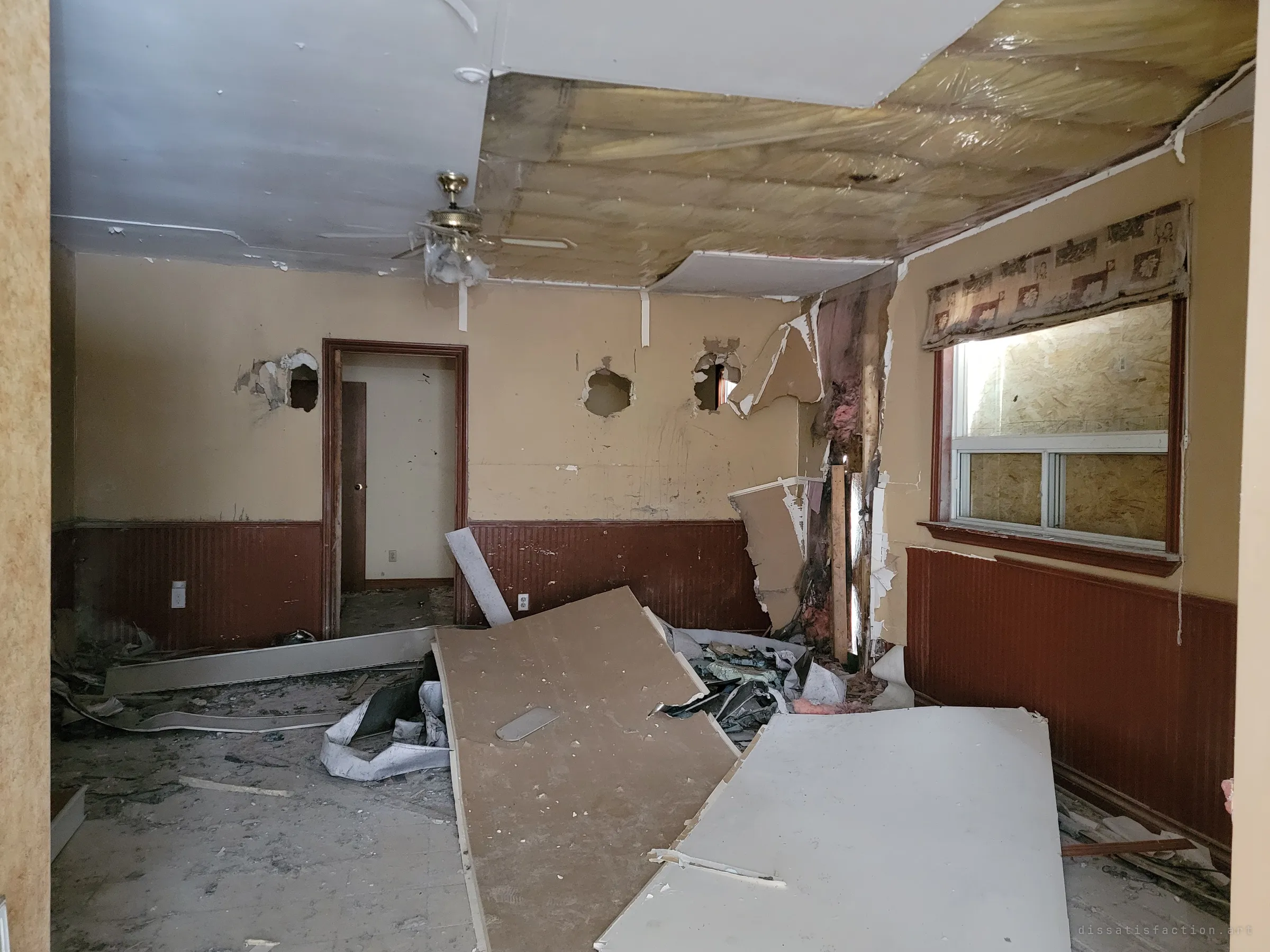 A living room half torn apart: ceiling panels hang down over exposed insulation, fist-sized holes punch the walls and drywall sheets lie across the floor. The window behind the valance is boarded from outside.