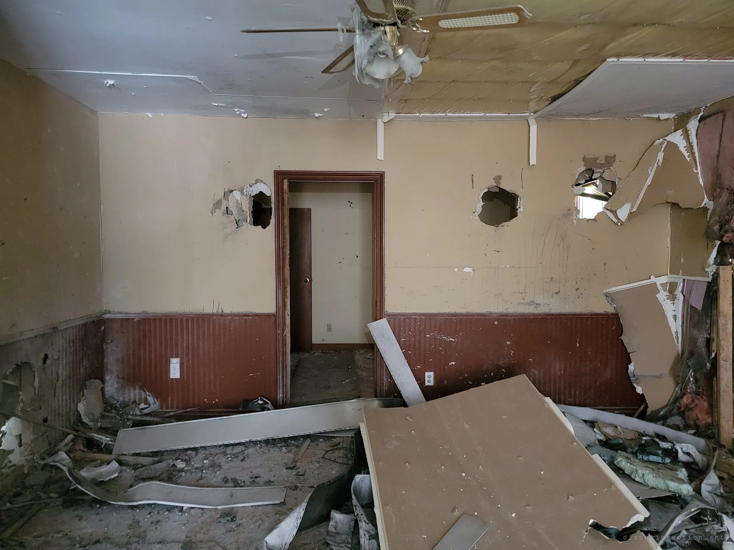 The same wrecked room seen straight on, a doorway framed by holes knocked through the plaster and a ceiling fan still hanging over fallen drywall and debris.