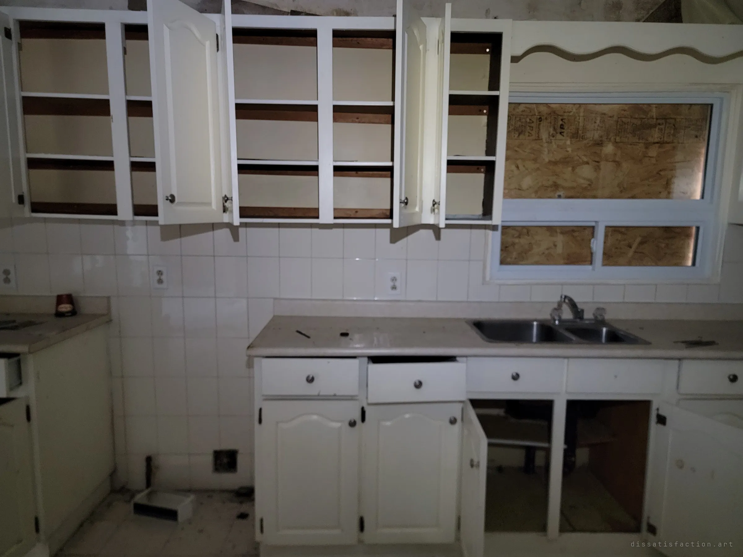 A dim kitchen with every cabinet door flung open and emptied, drawers pulled out and the window over the sink boarded with plywood.
